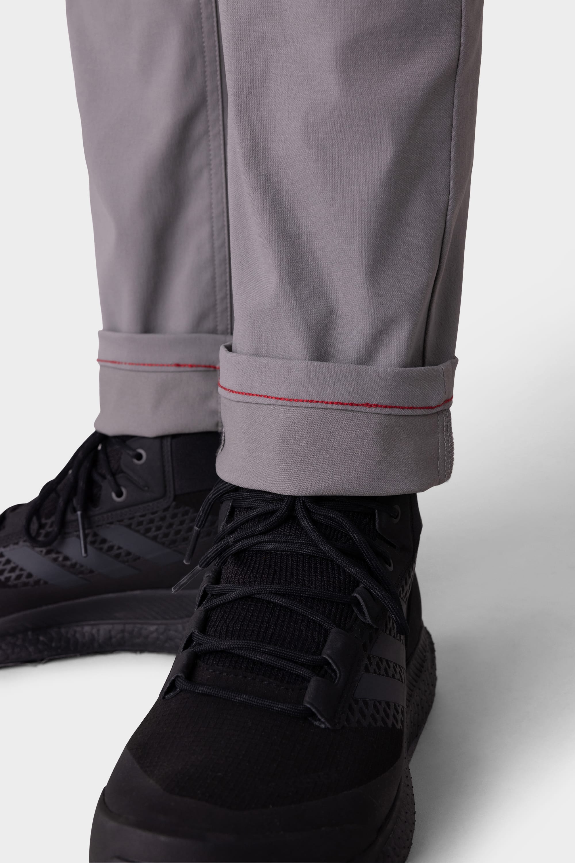 Close-up of a person wearing 686 Mens Everywhere Pant - Slim Fit in gray, featuring red stitch cuff detail and paired with black athletic shoes against a light background.