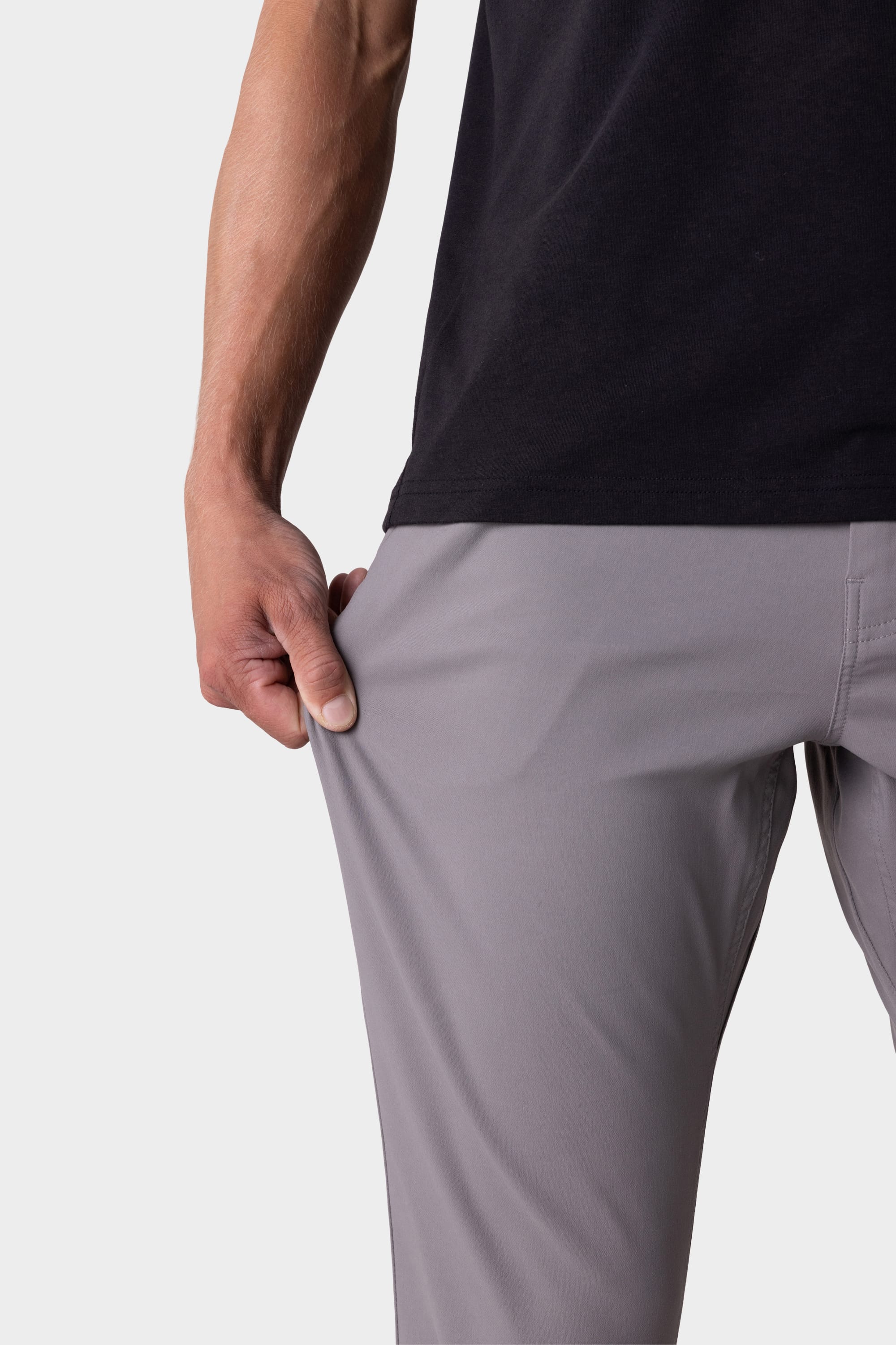 A man wearing the 686 Mens Everywhere Pant - Slim Fit in light gray and a black shirt stretches the fabric outward with one hand, showcasing the stretchiness and breathability of these 686 pants.