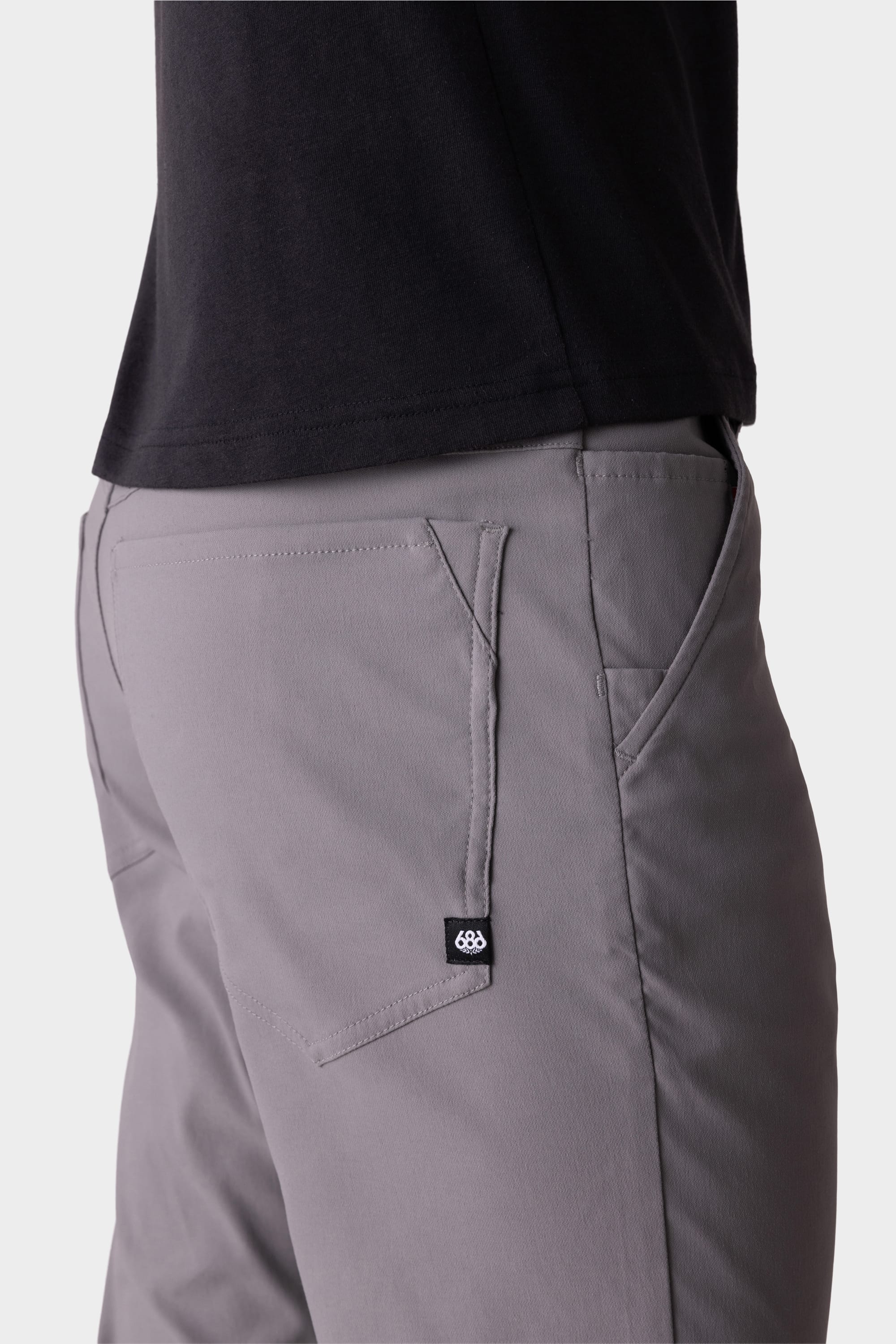 A person wears the 686 Mens Everywhere Pant - Slim Fit in light gray with a black tag and small white logo on the back pocket, paired with a black shirt. The image highlights the side and back of these stylish, breathable 686 pants.