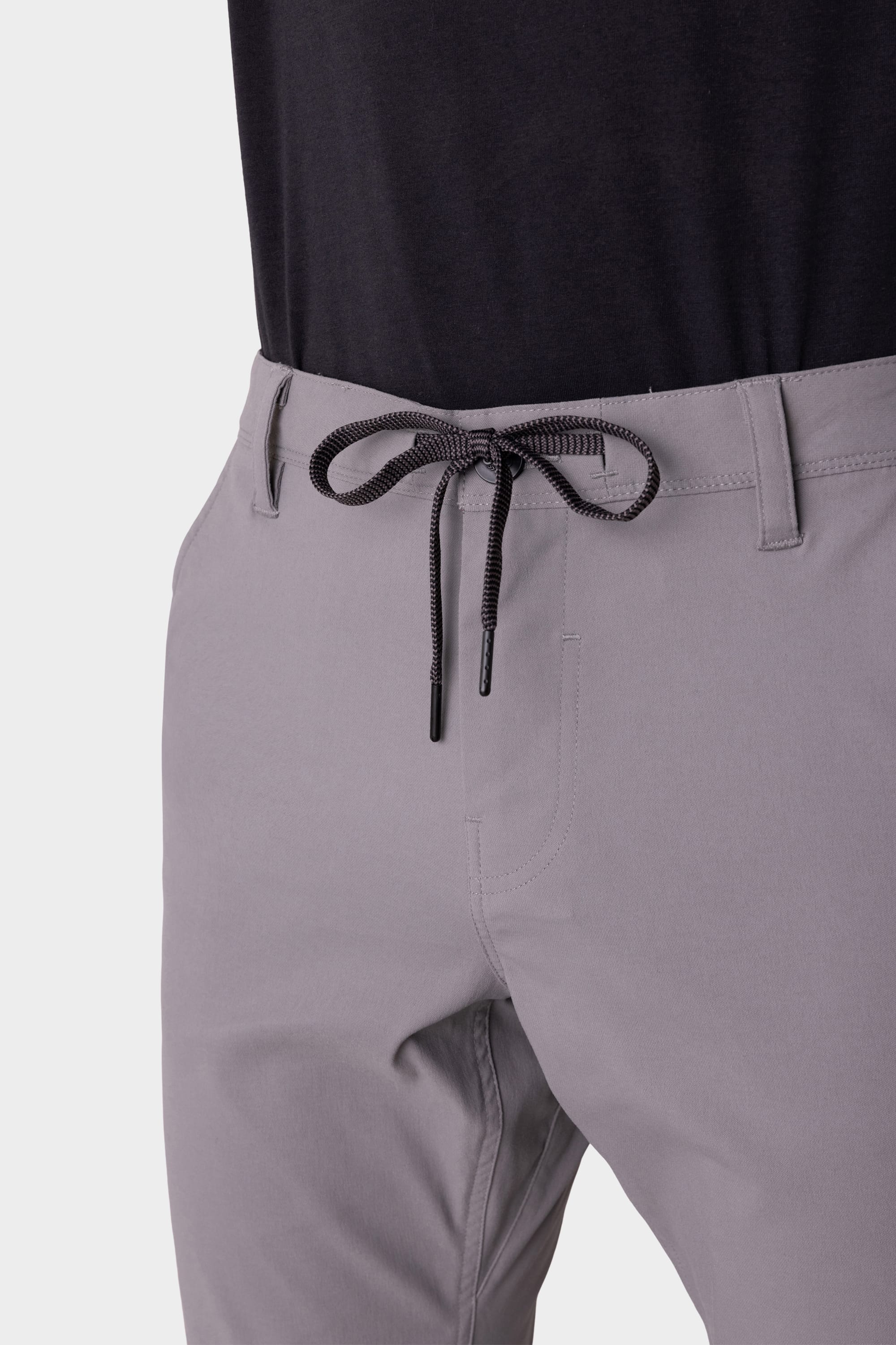 A person models the 686 Mens Everywhere Pant - Slim Fit in light gray, featuring a black and white drawstring waist, paired with a tucked-in black shirt. The photo displays these stain-resistant 686 pants from waist to mid-thigh.
