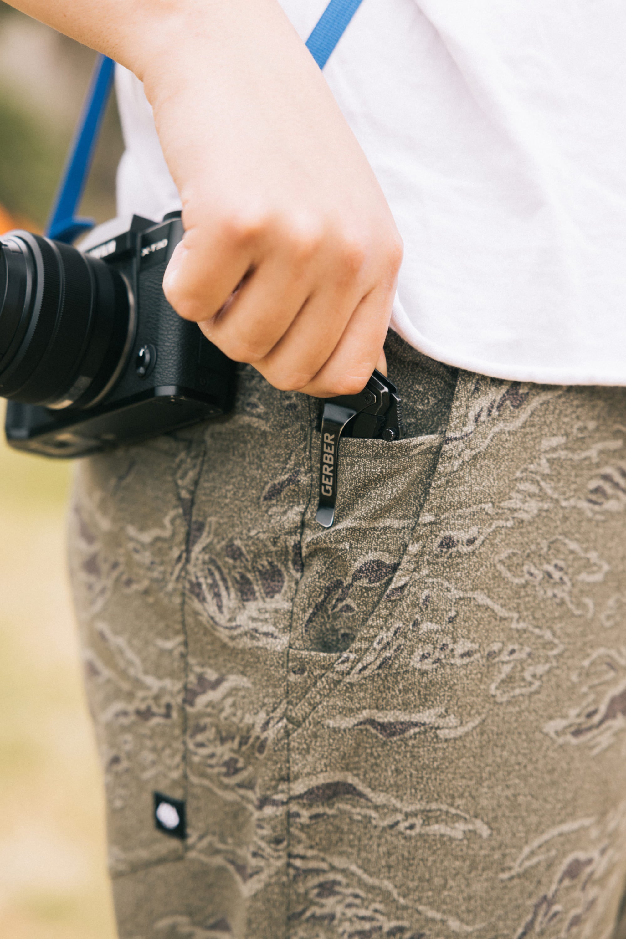 A person in 686 Men's Everywhere Pant - Slim Fit (camouflage, stain-resistant) holds a black multitool near their pocket, while a black camera with a blue strap hangs around their neck.