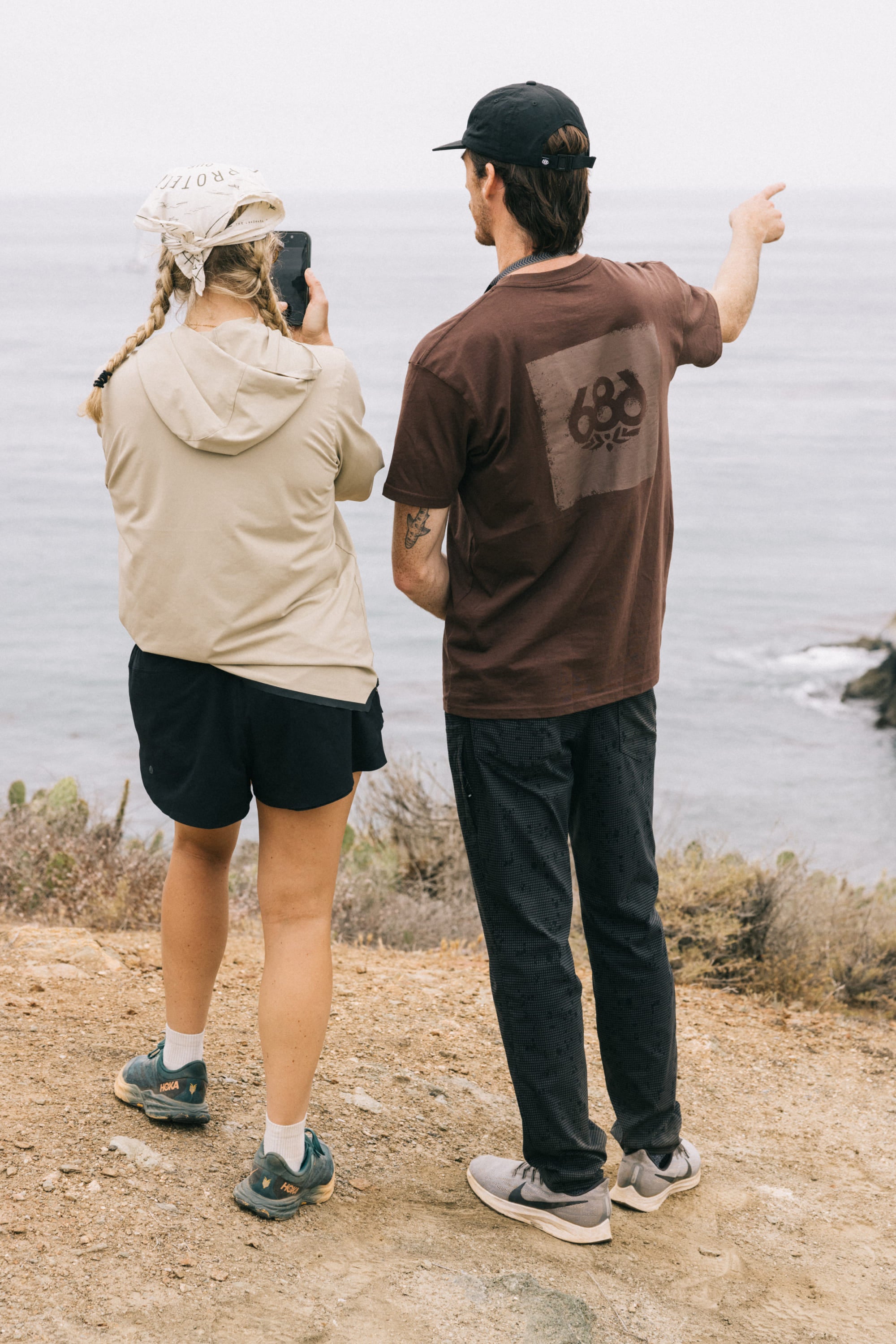 Two people stand on a rocky path by the ocean. One takes a photo while the other points toward the water. Both wear casual hats and 686 Men's Everywhere Pant - Slim Fit. Coastal plants and the sea appear in the background.