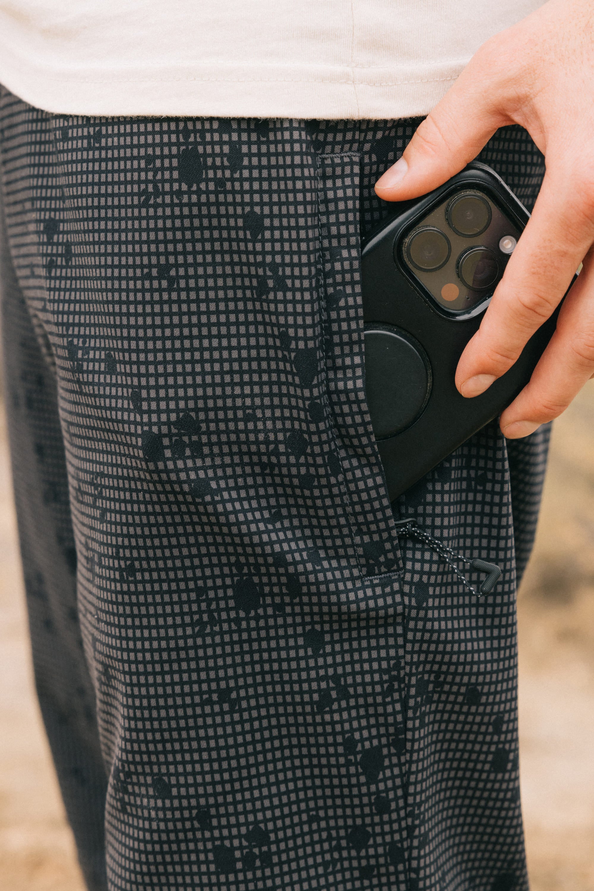 Someone slips a black smartphone with a camera bump into the pocket of 686 Men's Everywhere Pant - Slim Fit by 686, featuring a dark pattern and stain-resistant fabric, while the lower part of a light shirt is visible.