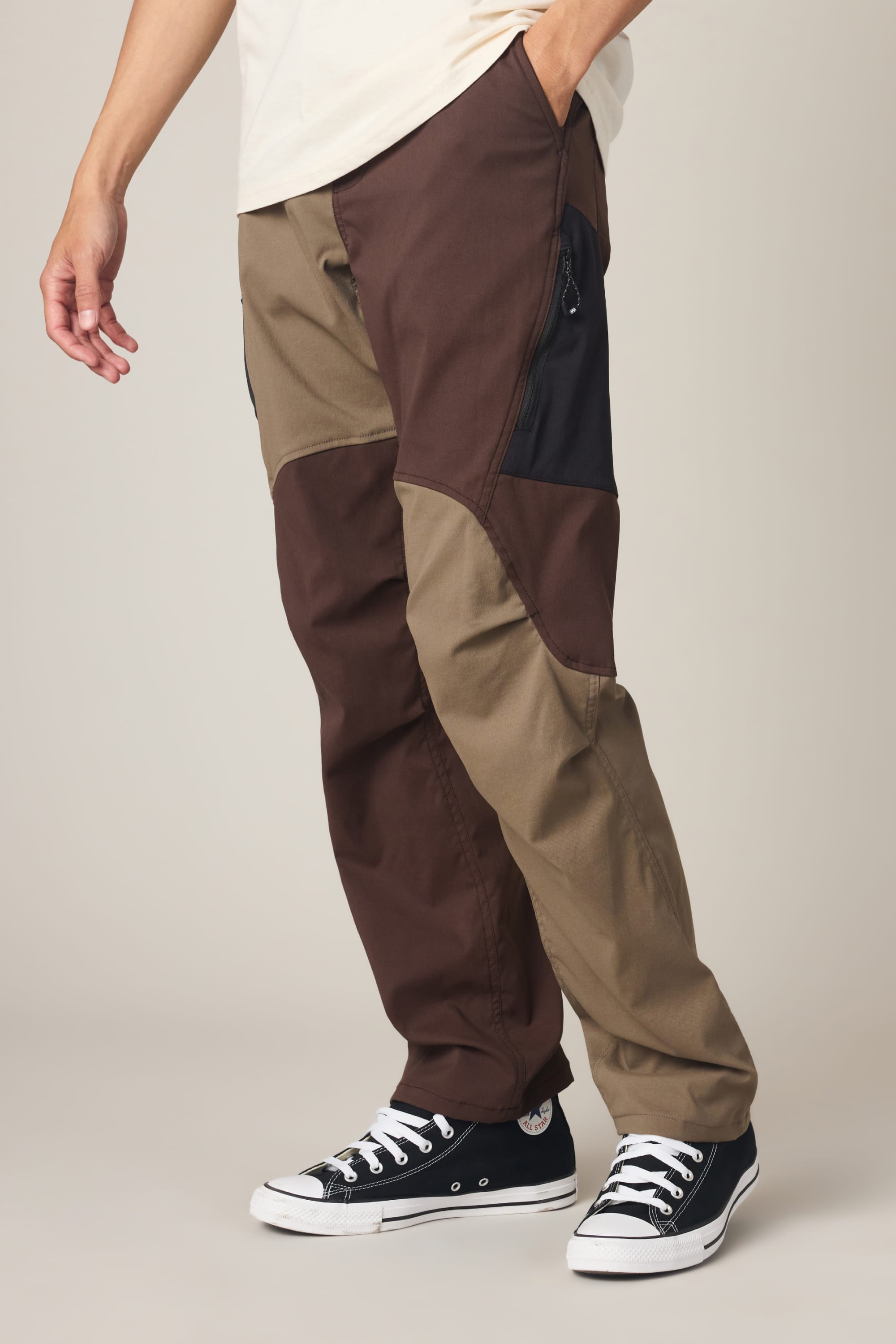 A person wearing the 686 Men's Anything Cargo Pant - Relaxed Fit by 686, featuring a color-blocked brown and tan design with black patches, 4-way stretch fabric, and a 13-pocket layout, stands with one hand in their pocket and pairs the pants with black low-top Converse sneakers.