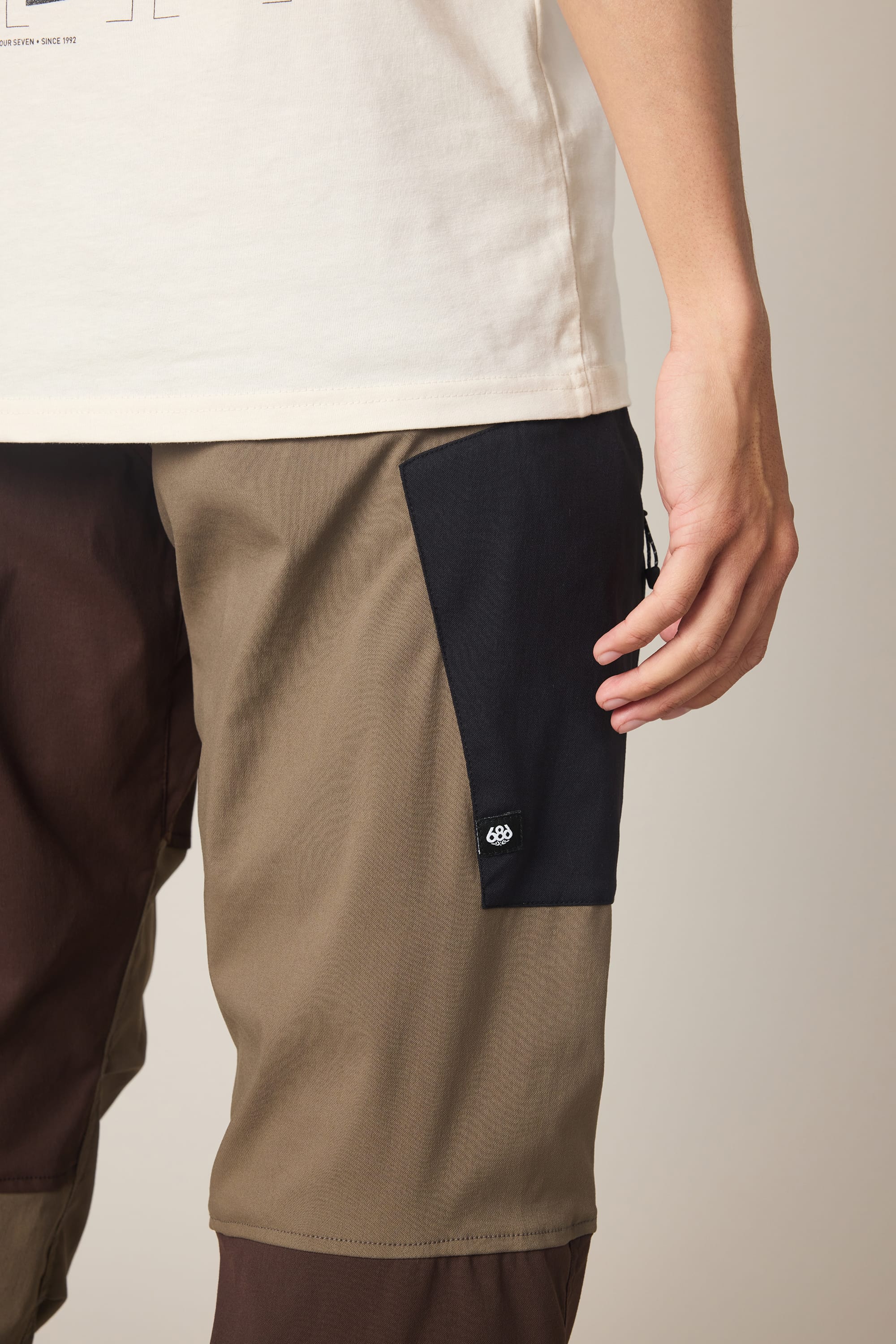 A person wearing the 686 Men's Anything Cargo Pant - Relaxed Fit in a tan and brown color-block design, featuring a black pocket patch and technical cargo pant styling, stands with one hand relaxed at their side, partially visible against a neutral background.