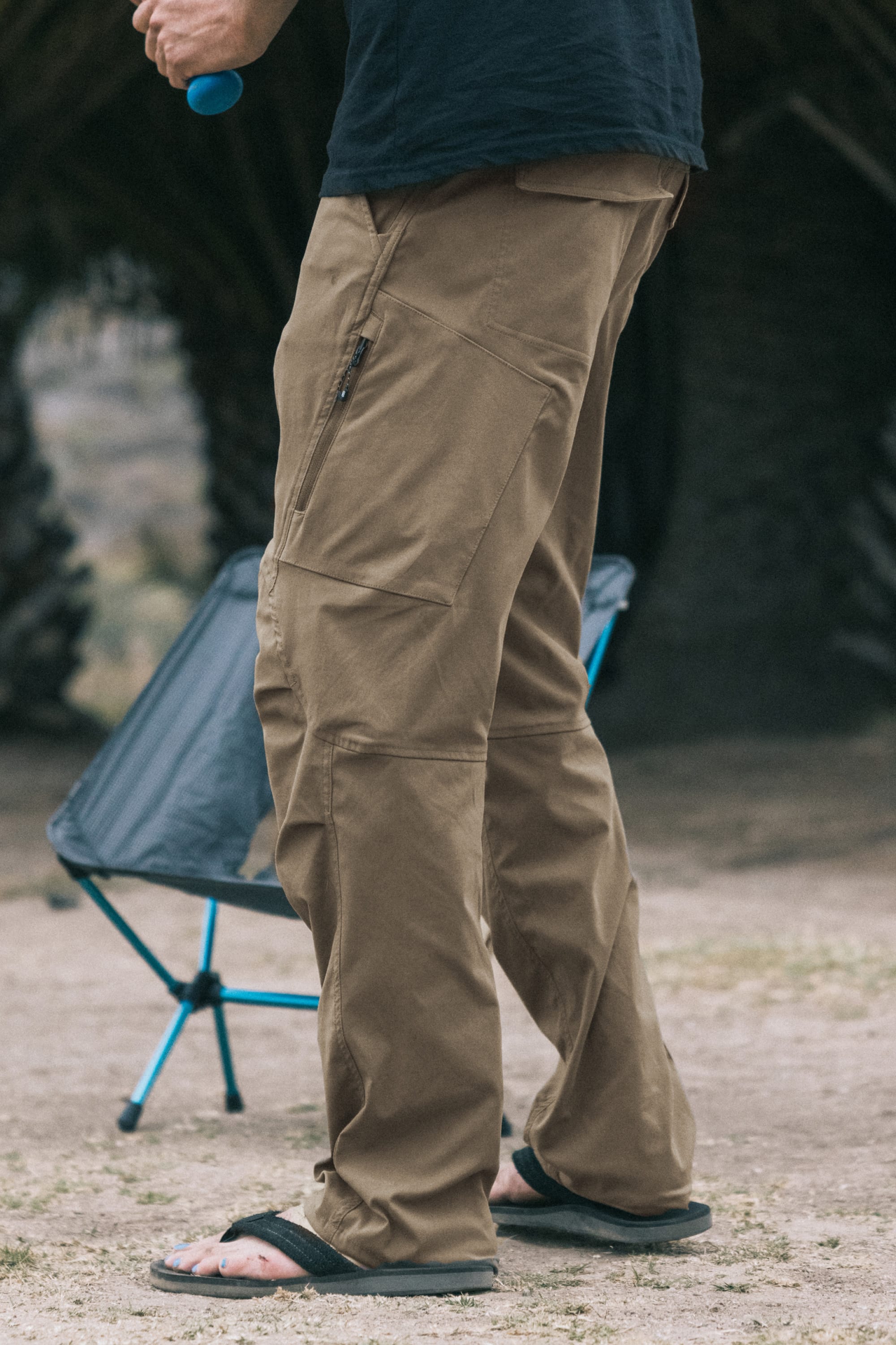 A person wearing 686 Men's Anything Cargo Pant - Relaxed Fit in tan with a black shirt and sandals stands outside by a small blue folding chair, holding a blue item. Trees and blurred scenery create a laid-back atmosphere.