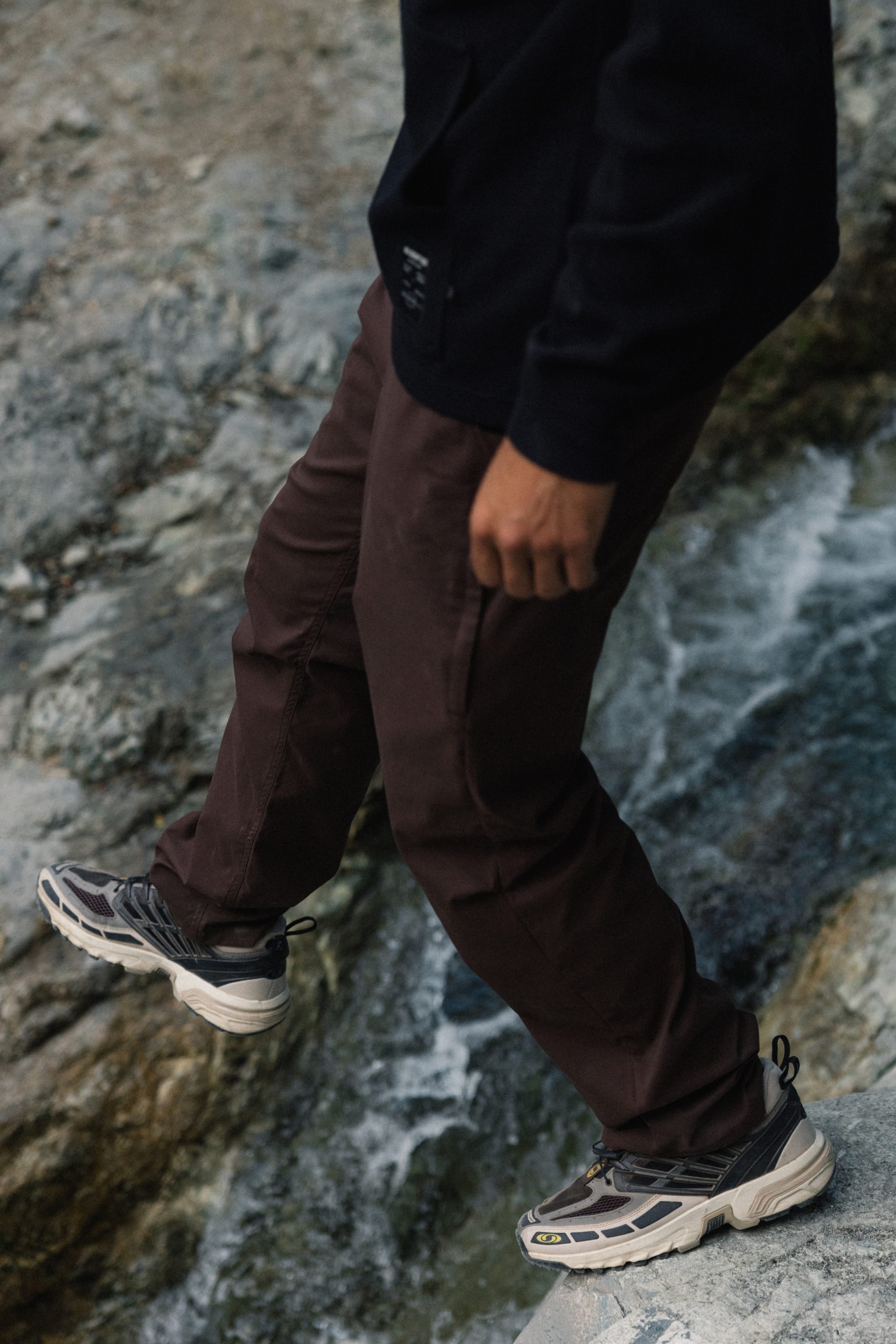 Wearing the 686 Men's Everywhere® Pant - Relaxed Fit by 686 in brown, a person strolls across rocky terrain near flowing water, paired with a black sweatshirt and gray sneakers.