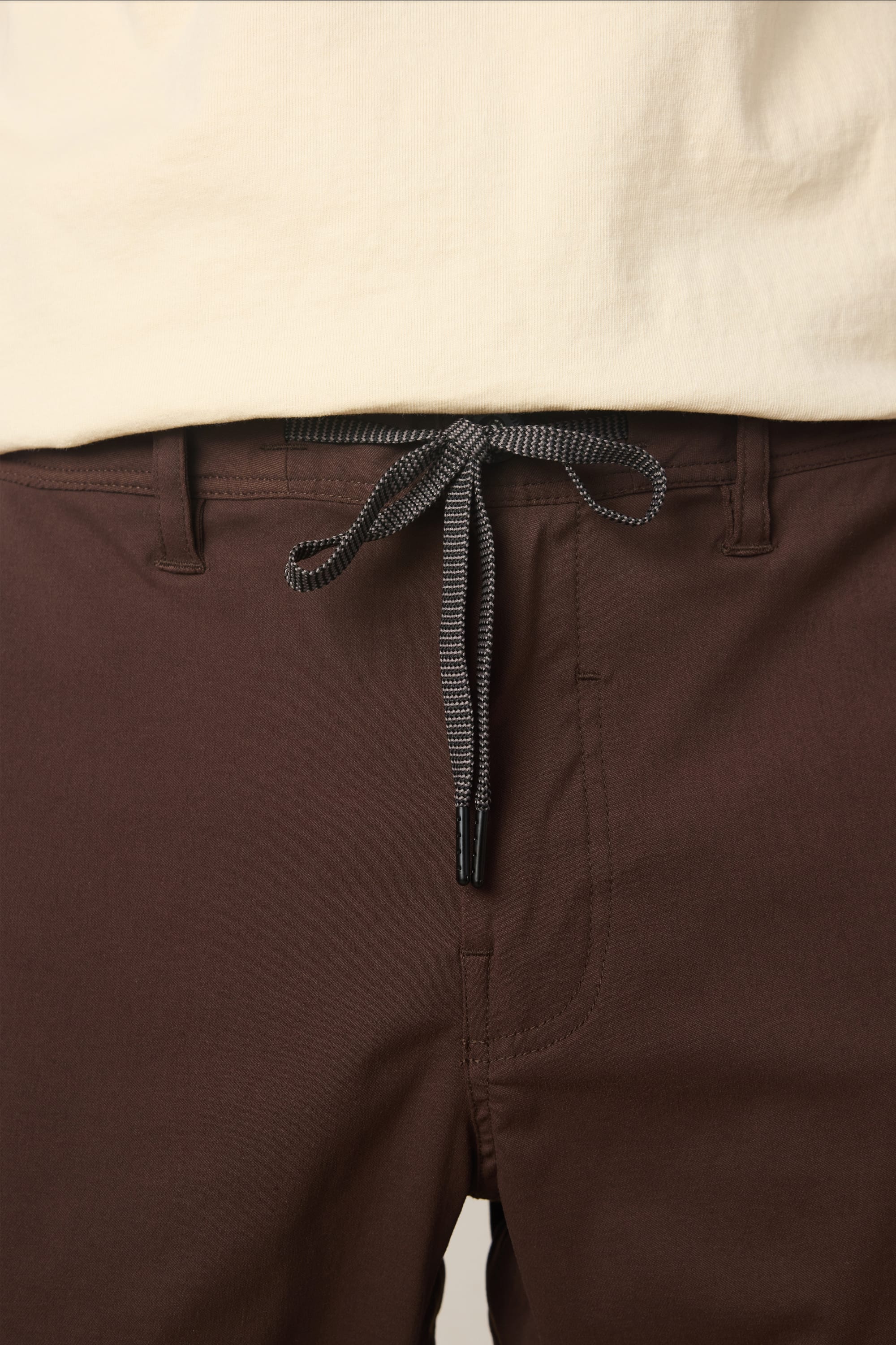 A close-up of a person wearing the 686 Men's Everywhere® Pant - Relaxed Fit in brown, featuring a black-and-white drawstring, paired with a cream-colored shirt.