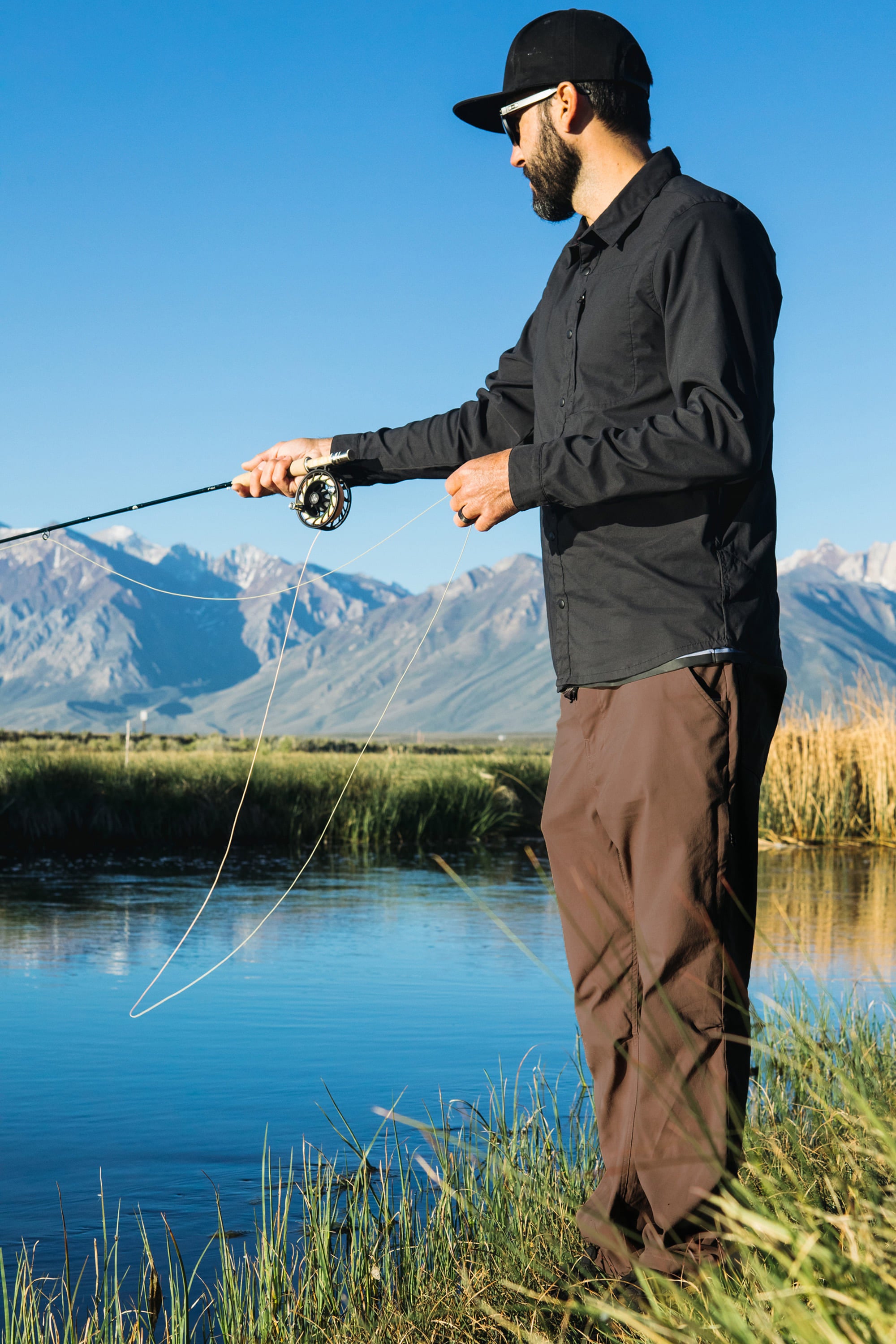A man wearing a black shirt, 686 Men's Everywhere® Pant - Relaxed Fit, and a cap is fly fishing by a river with mountains and a clear blue sky in the background.