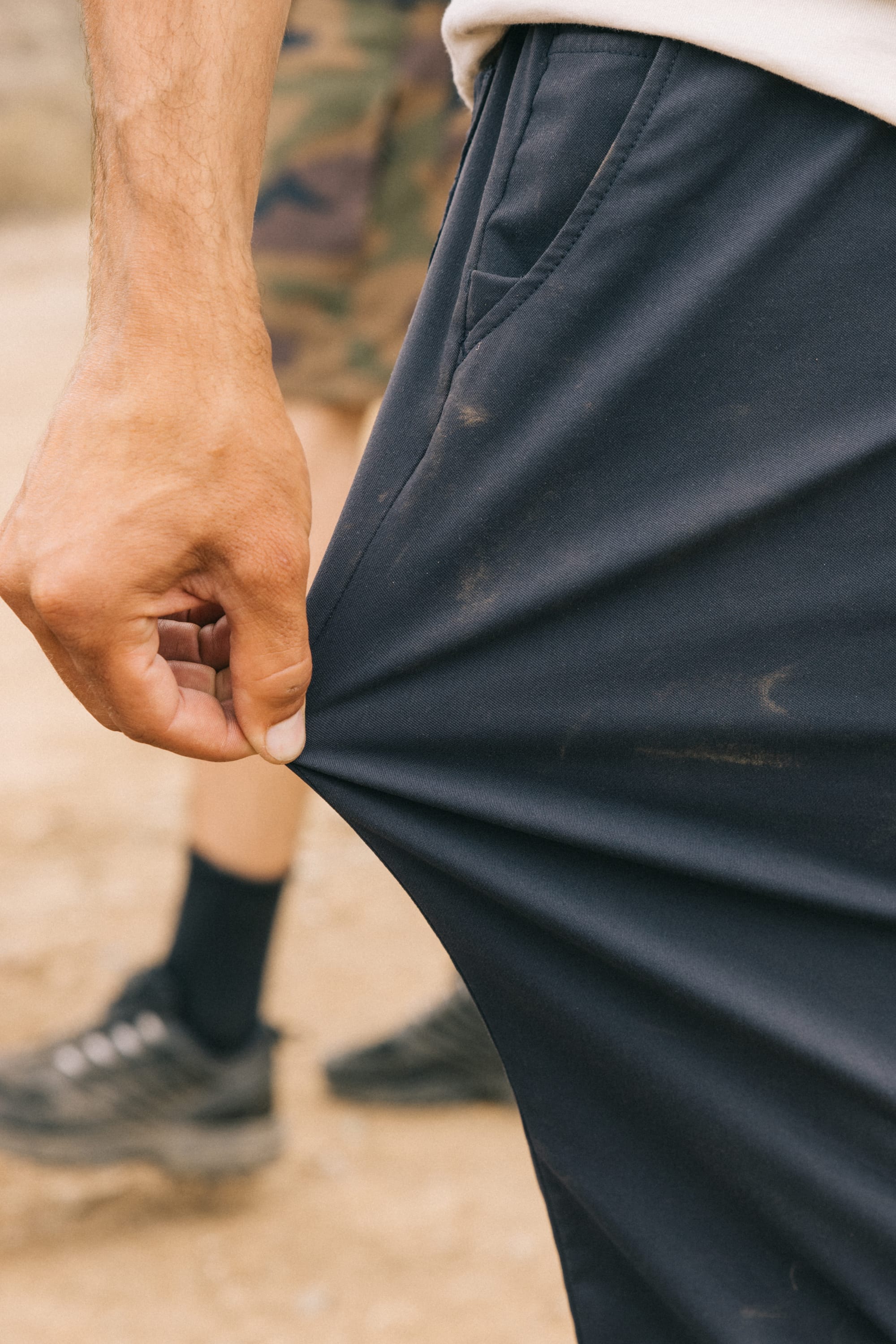 A person outdoors pulls the fabric of their 686 Men's Everywhere® Pant - Relaxed Fit. In the blurred background, another individual in camouflage shorts and black shoes stands.