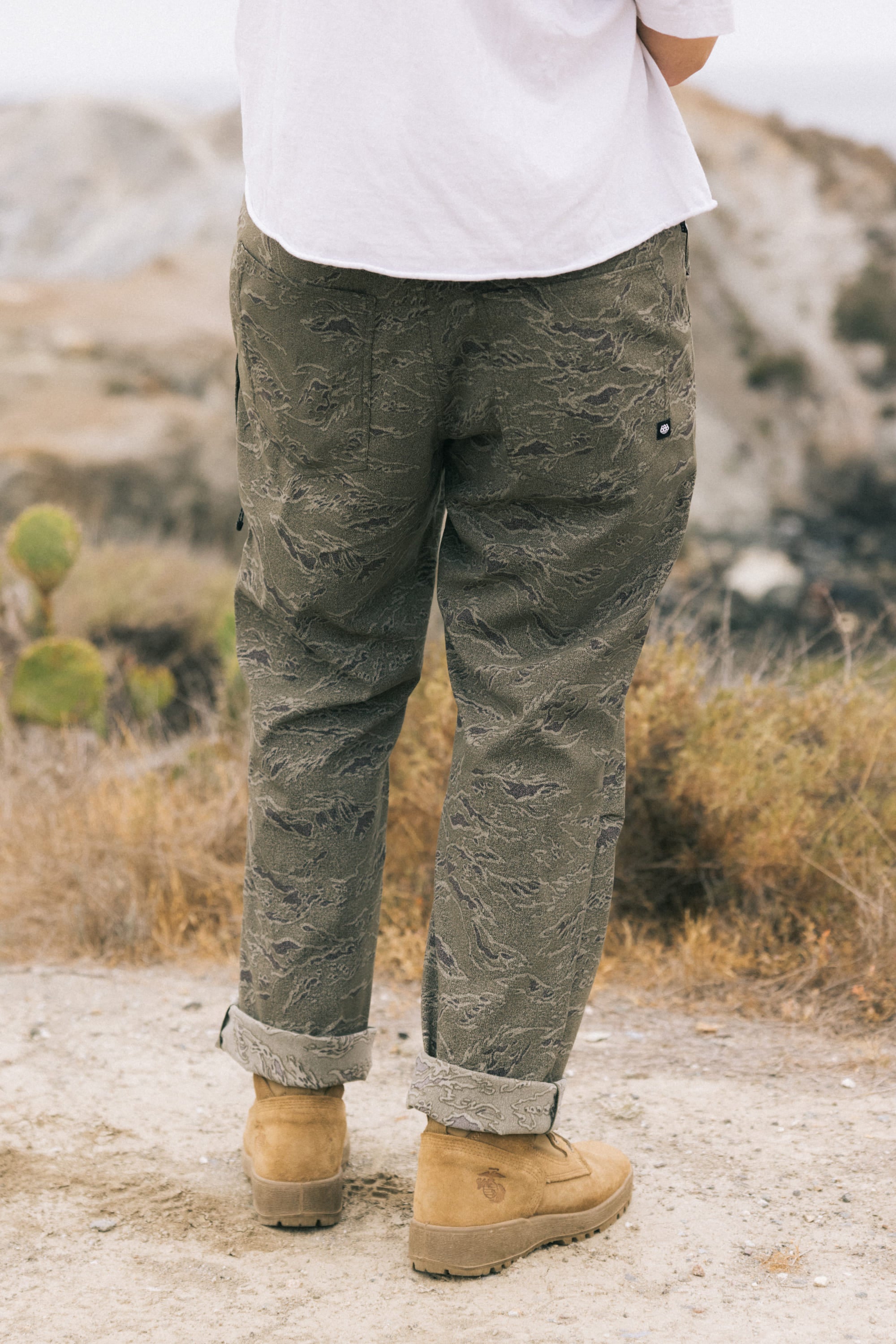 A person stands outdoors on a dirt path, facing away, wearing 686 Men's Everywhere Pant - Relaxed Fit in olive green, a white shirt, and tan suede boots. The background features dry grass, rocks, and a blurred landscape.
