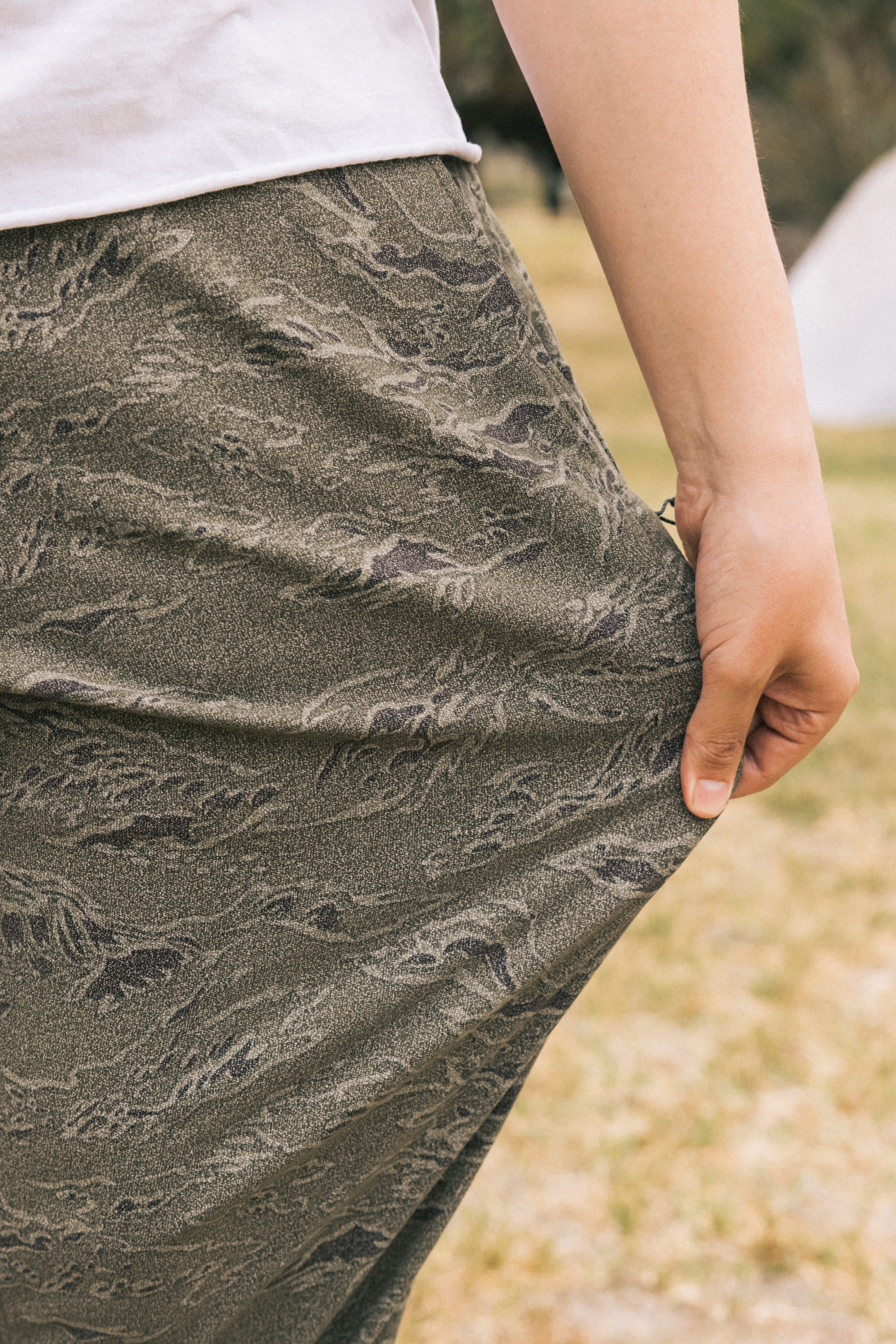 A person stands outdoors in grass and greenery, wearing 686 Men's Everywhere Pant - Relaxed Fit in green camo and a white shirt, holding a handful of the pant's fabric at their side.