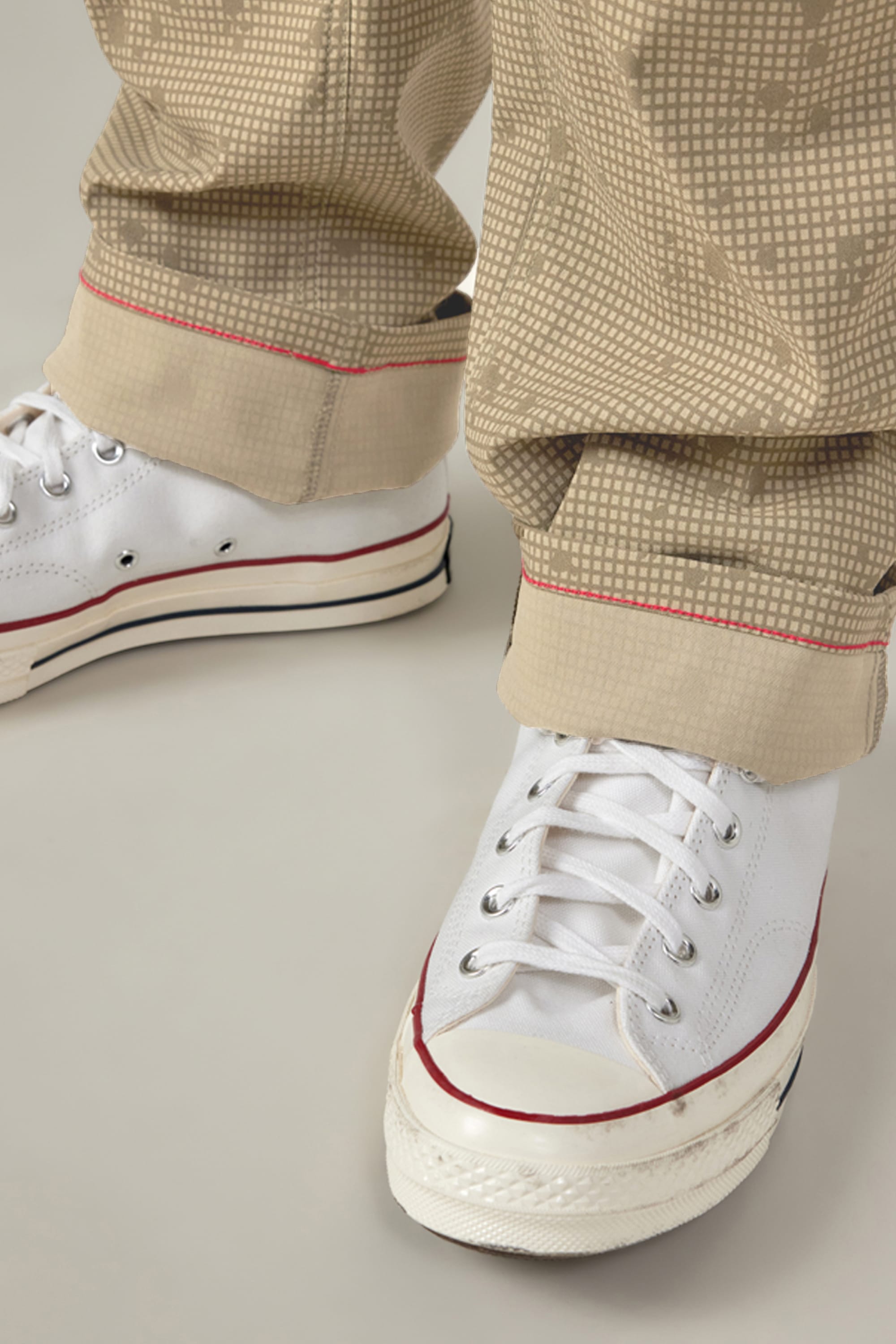 Close-up of a person wearing white lace-up sneakers and 686 Men's Everywhere® Pant - Relaxed Fit in beige with a grid pattern and red stitched rolled cuffs, blending comfort and style from 686.