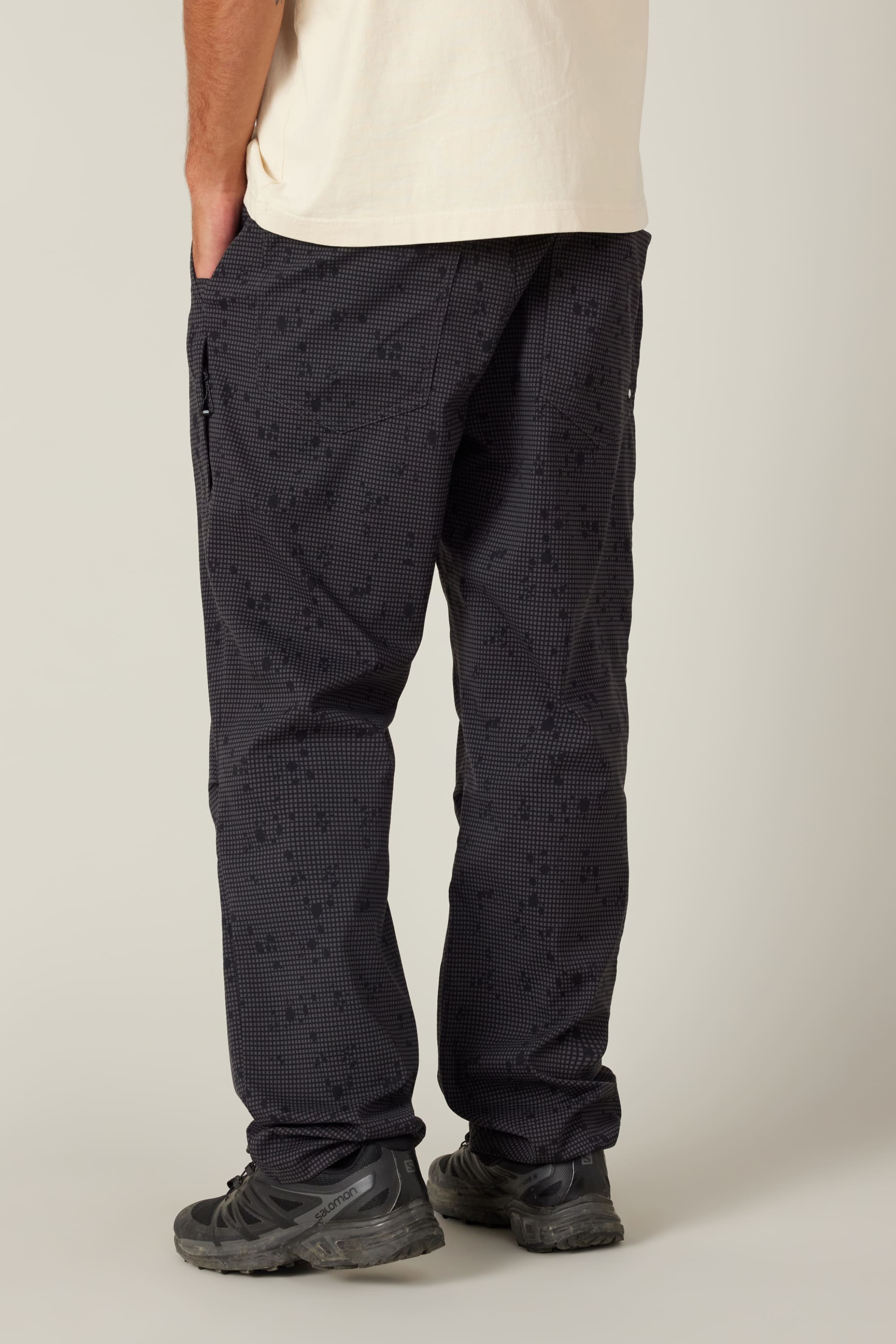 A person stands with their back to the camera, wearing a cream short-sleeve shirt and 686 Men's Everywhere Pant - Relaxed Fit by 686, paired with dark trail shoes. Their left hand is in their pocket against a plain light background.