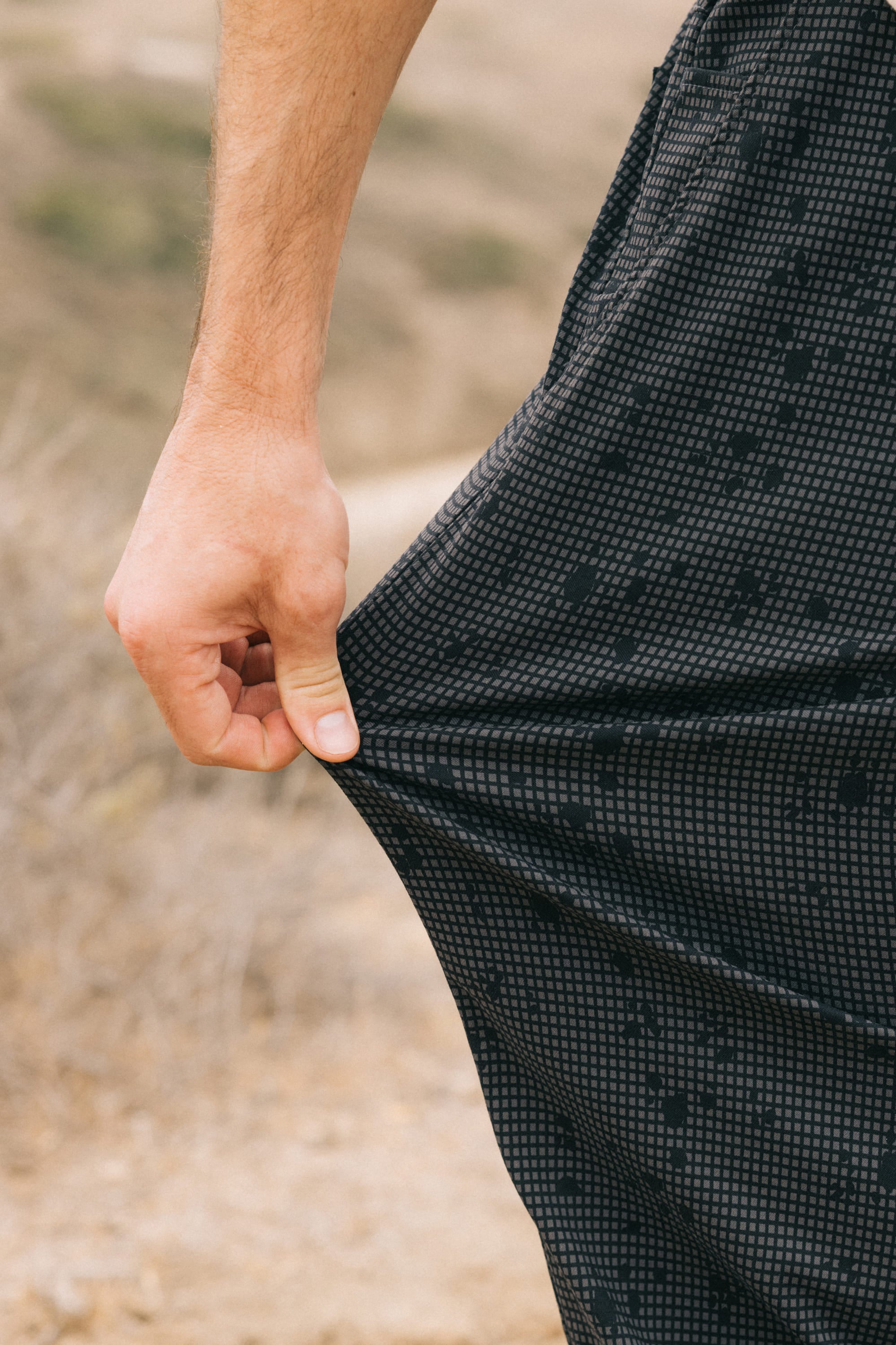 A hand stretches a section of the 686 Men's Everywhere Pant - Relaxed Fit, featuring a black breathable fabric with a small grid pattern, outdoors against a blurry, dry landscape.