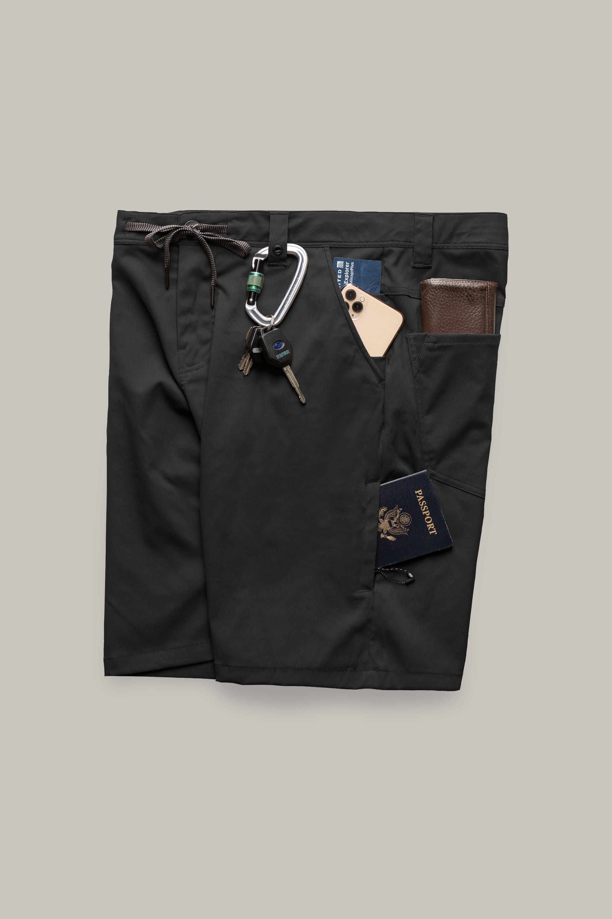 A pair of 686 Men's Everywhere® Hybrid Shorts in black, featuring multiple pockets holding a carabiner with keys, a pen, phone, wallet, and blue passport, made from breathable, stain-resistant fabric on a light gray background.