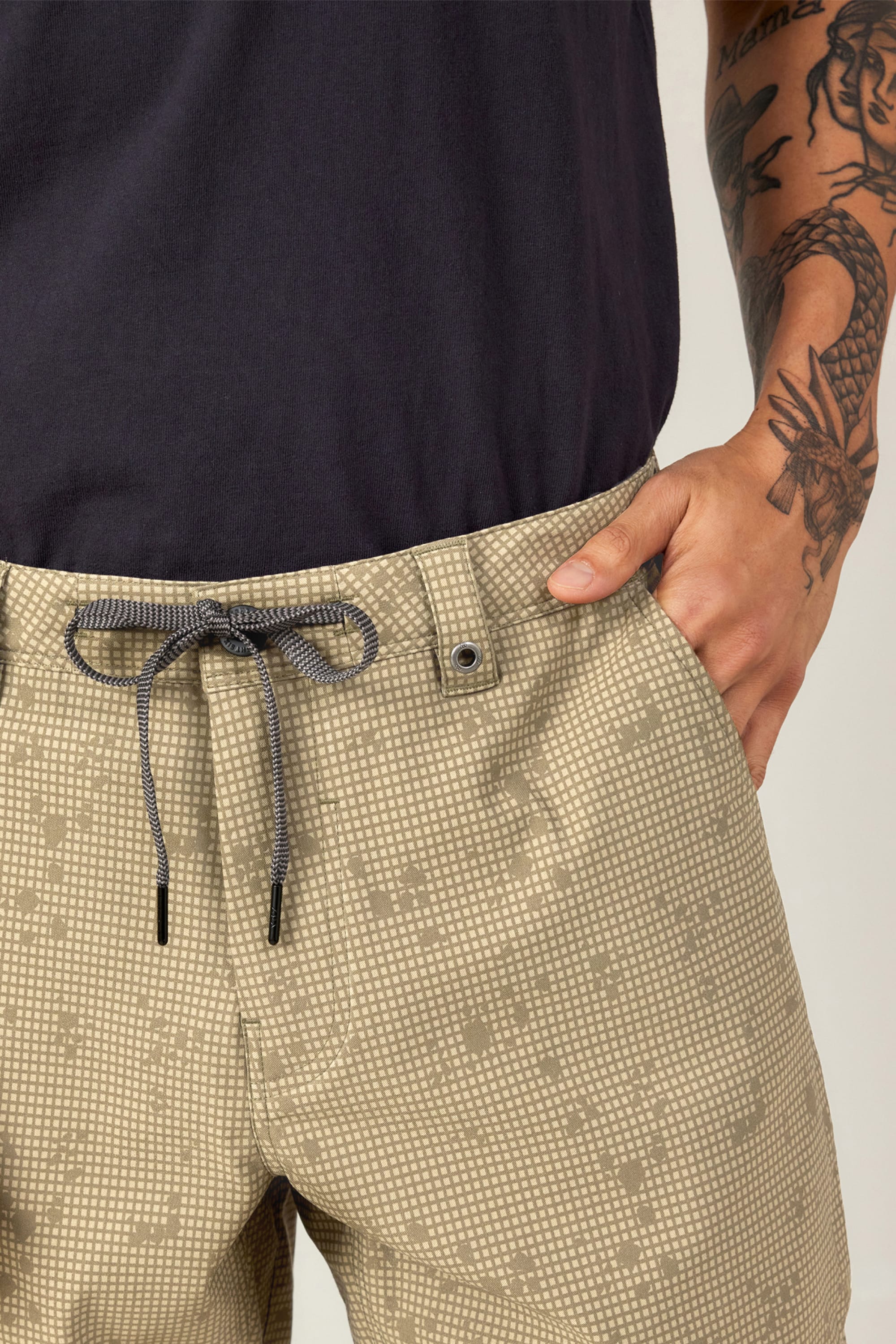 A person wears 686 Men's Everywhere® Hybrid Short in light tan with a pattern, drawstring, and one hand in the pocket, paired with a black shirt. A dragon tattoo is visible on their arm.