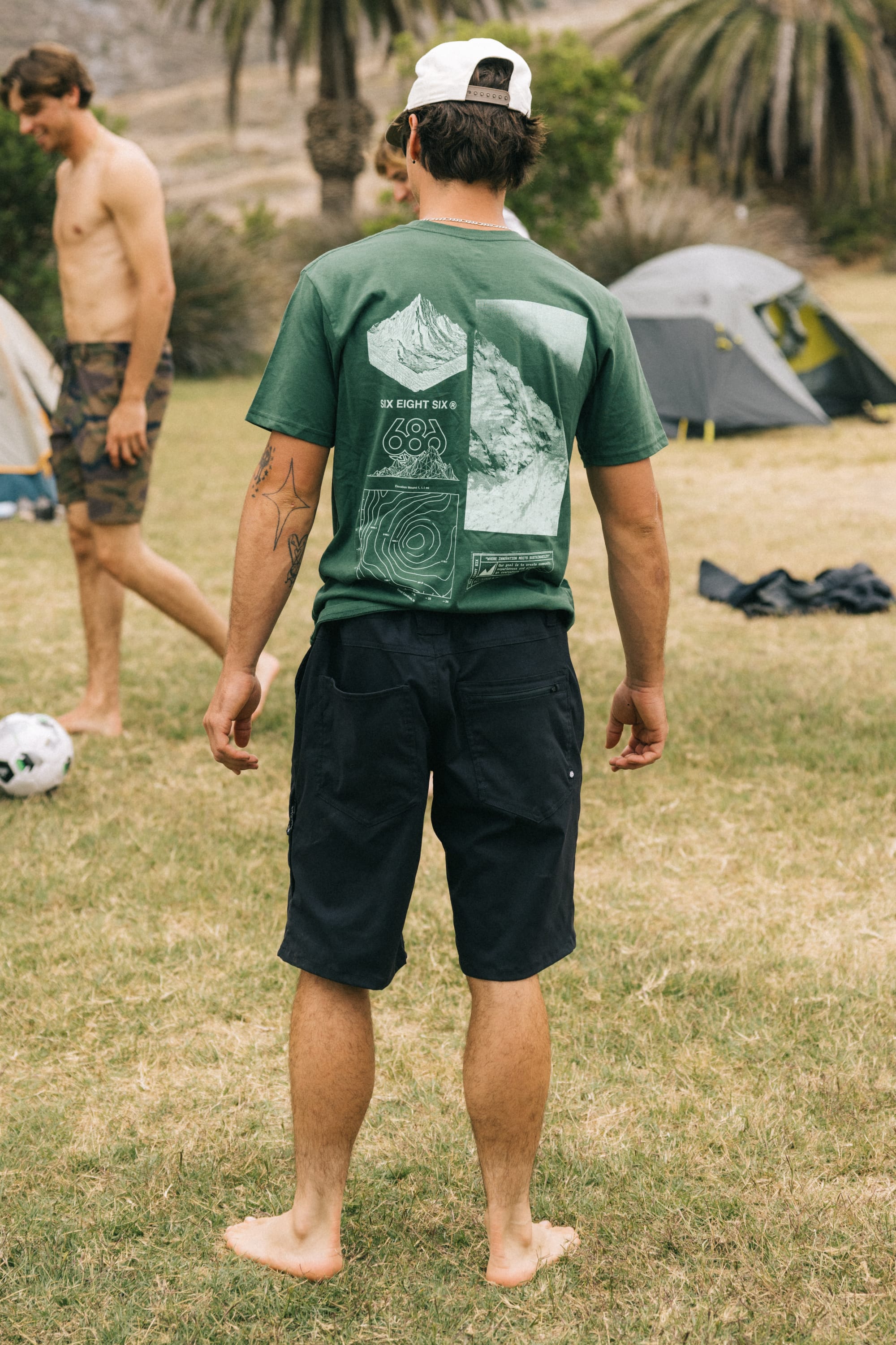A man stands barefoot on grass, facing away, wearing a breathable green graphic tee, a white cap, and 686 Men's Everywhere® Hybrid Short - Relaxed Fit by 686. Behind him are another man, tents, and palm trees at a campsite.
