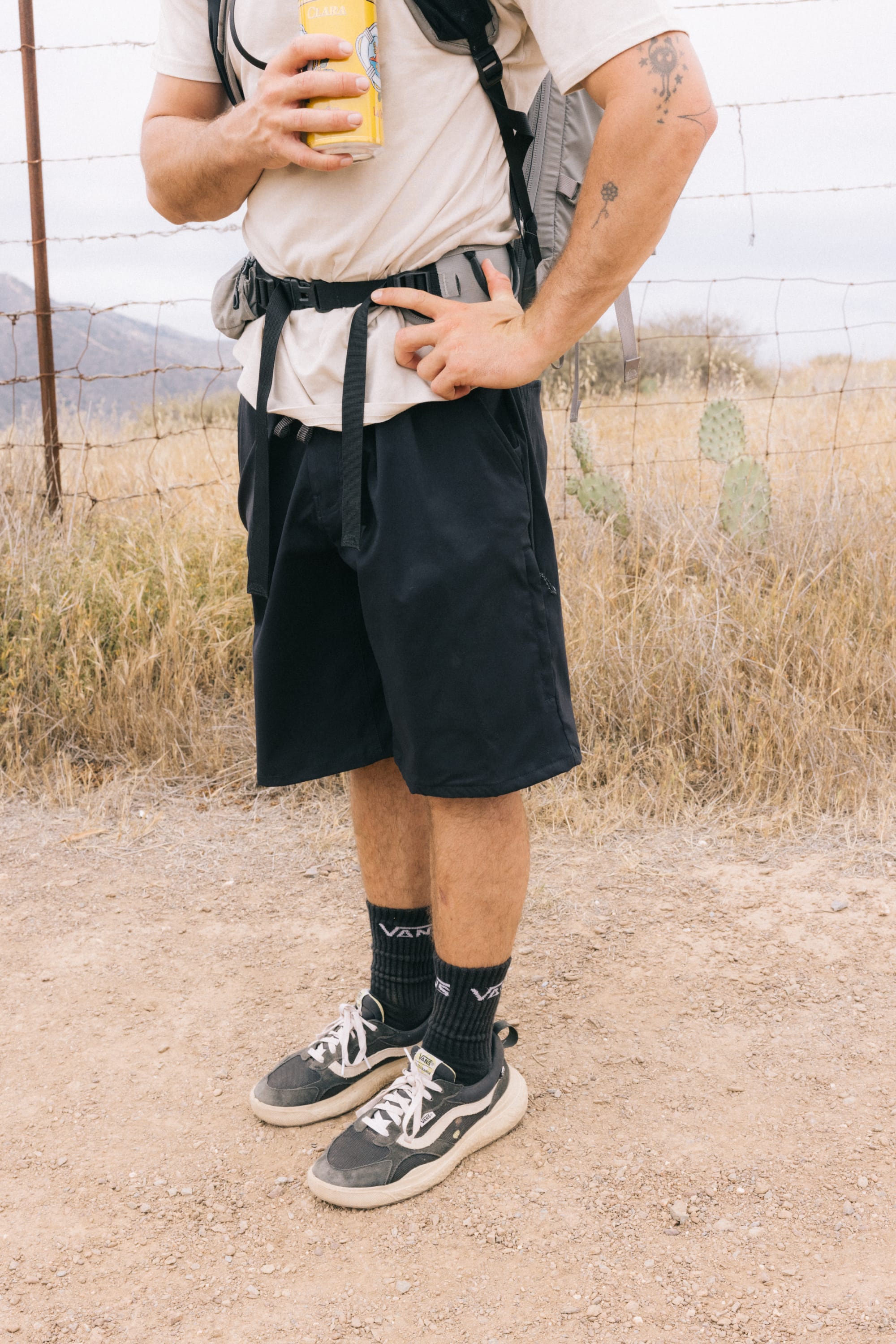 A person stands on a dirt path wearing 686 Men's Everywhere® Hybrid Short - Relaxed Fit, black socks, sneakers, a beige shirt, and backpack. They hold a can amid dry grass, cacti, and a wire fence in the background.