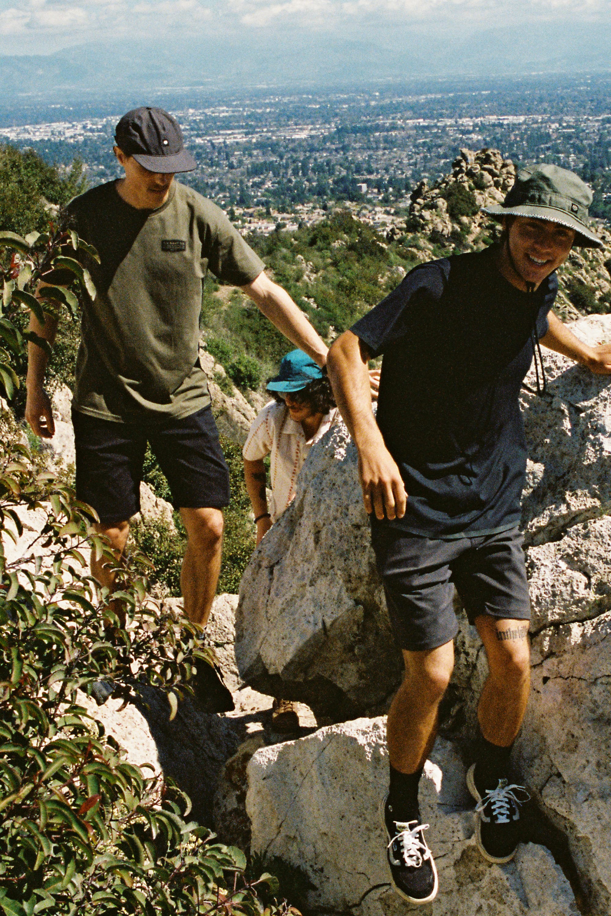 Three people wearing 686 Men's Everywhere® Hybrid Short - Relaxed Fit by 686 hike over rocks on a hillside in casual, breathable style, with a cityscape and clear sky in the background.