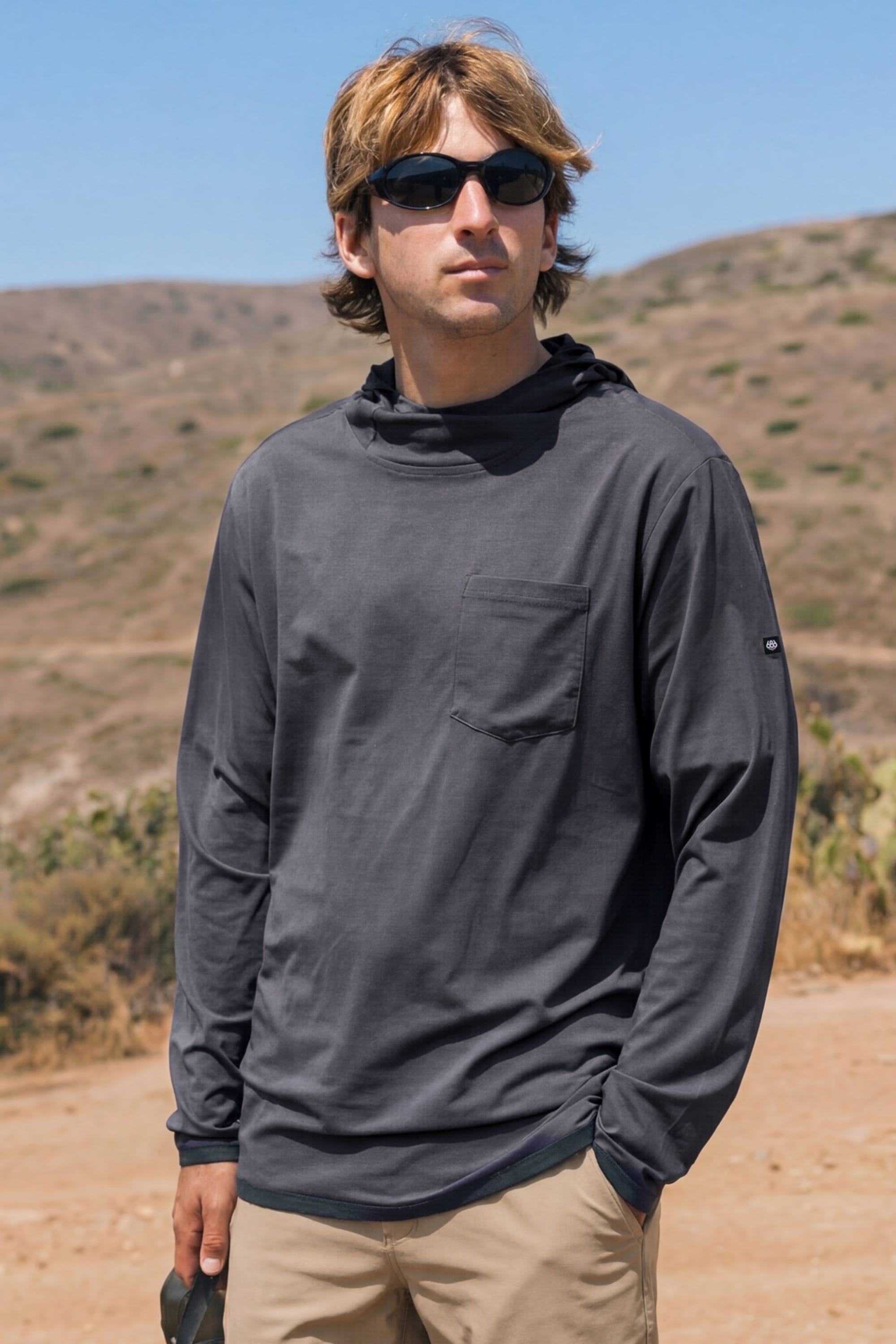 Wearing sunglasses, the person stands outdoors in light-colored pants and the 686 Men's Let's Go Tech Hoody by 686, which features a chest pocket and sun protection, with sunny hills and sparse vegetation in the background.