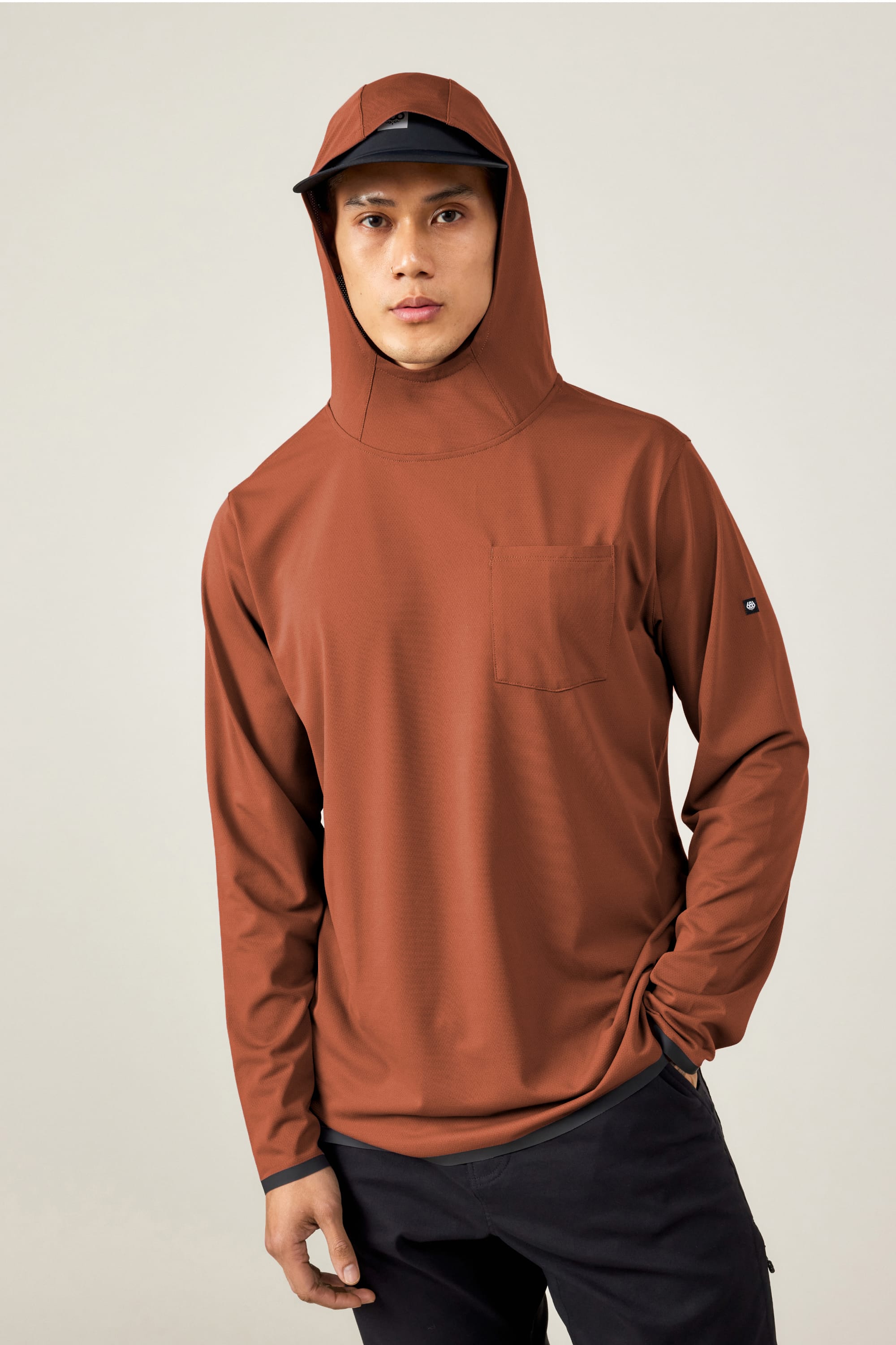 A person in the 686 Men's Let's Go Tech Hoody in rust and a black cap stands against a plain light background.