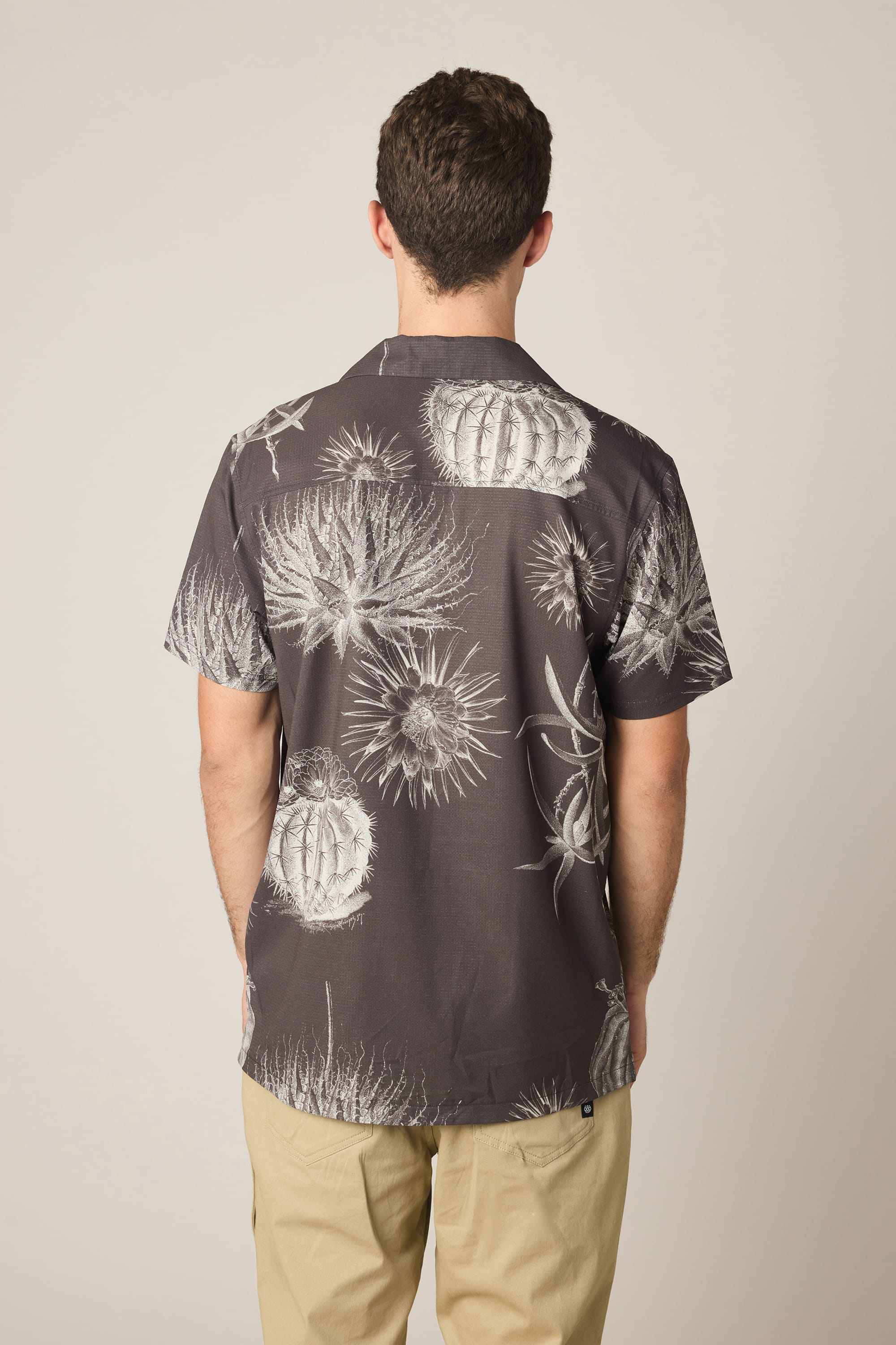 A man, seen from behind against a plain background, wears the 686 Men's Nomad Perforated Button Down Shirt by 686—a dark lightweight shirt featuring a white cactus print—paired with light-colored pants.