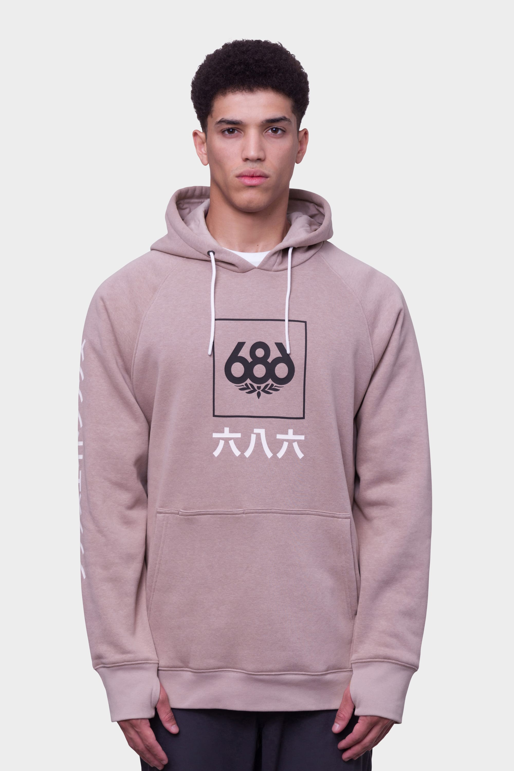 A young man wears the 686 Mens Script Knockout Hoody, featuring a beige color, black and white 686 logo with Asian characters on the front, white drawstrings, and a kangaroo pocket, standing against a plain white background.
