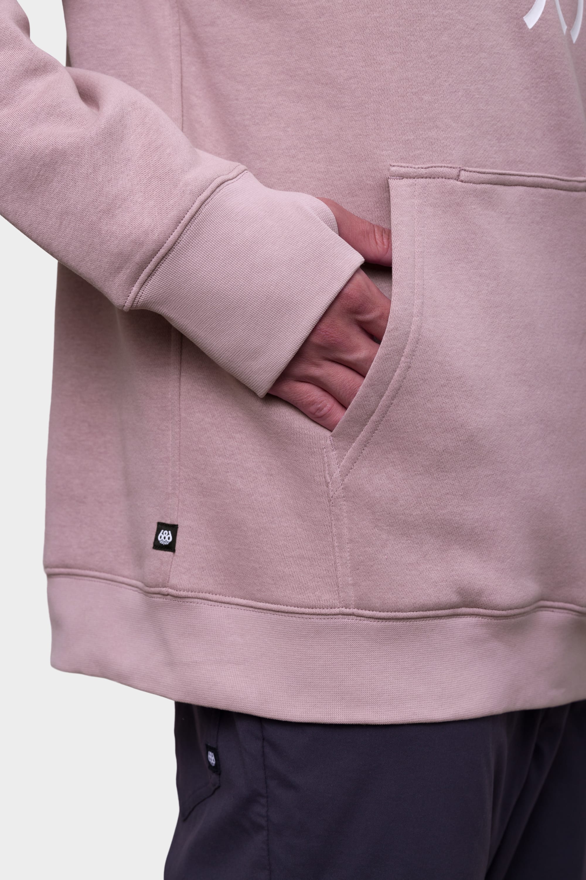 A person wears the 686 Mens Script Knockout Hoody in light pink, with their right hand in the front pocket. The hoody features a small black tag near the pocket and is paired with dark pants against a plain, light background.