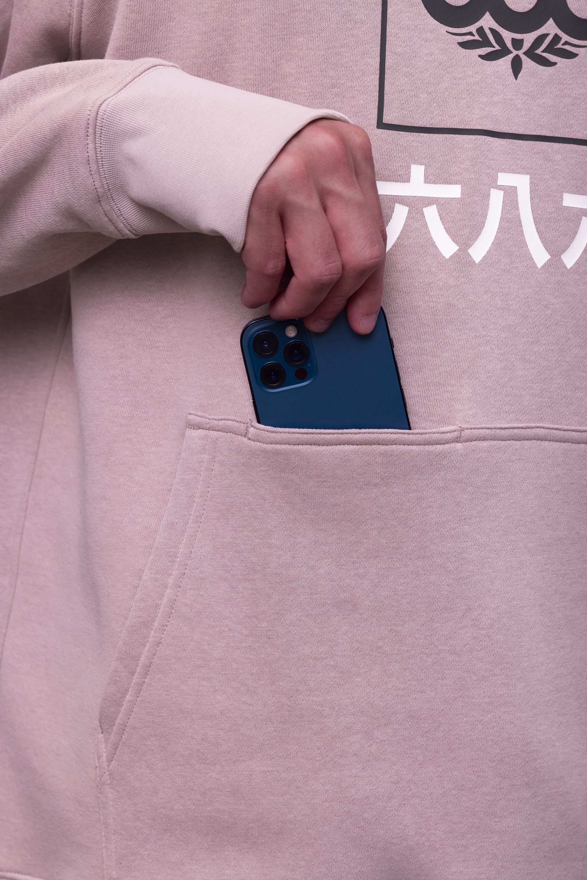 A person wearing a 686 Mens Script Knockout Hoody in beige places a blue smartphone into the front pocket, highlighting the hand, pocket, and part of the hoodie’s unique design.