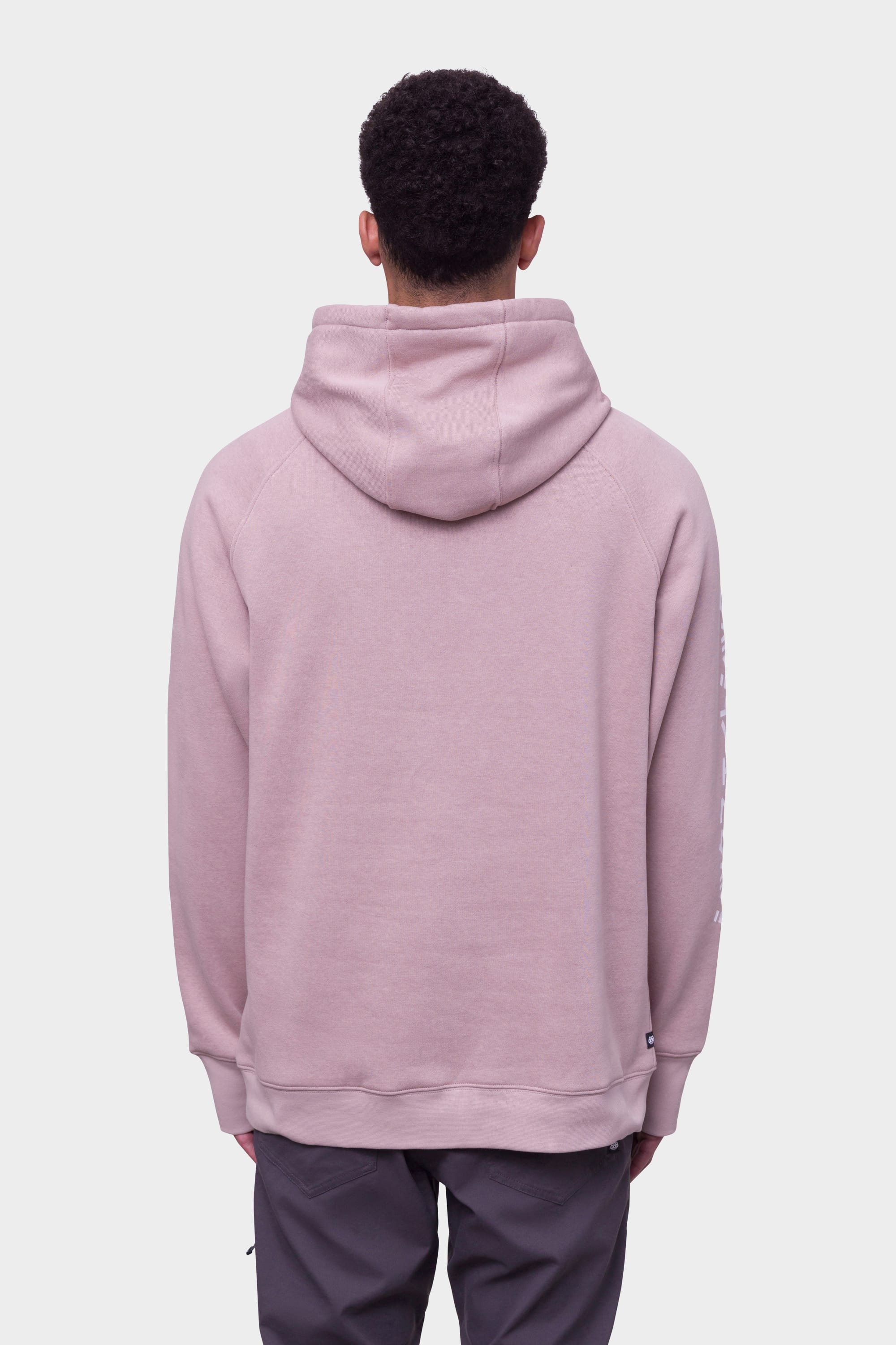 A person with short curly hair is seen from behind wearing the 686 Mens Script Knockout Hoody in light pink and dark pants, standing against a plain white background.