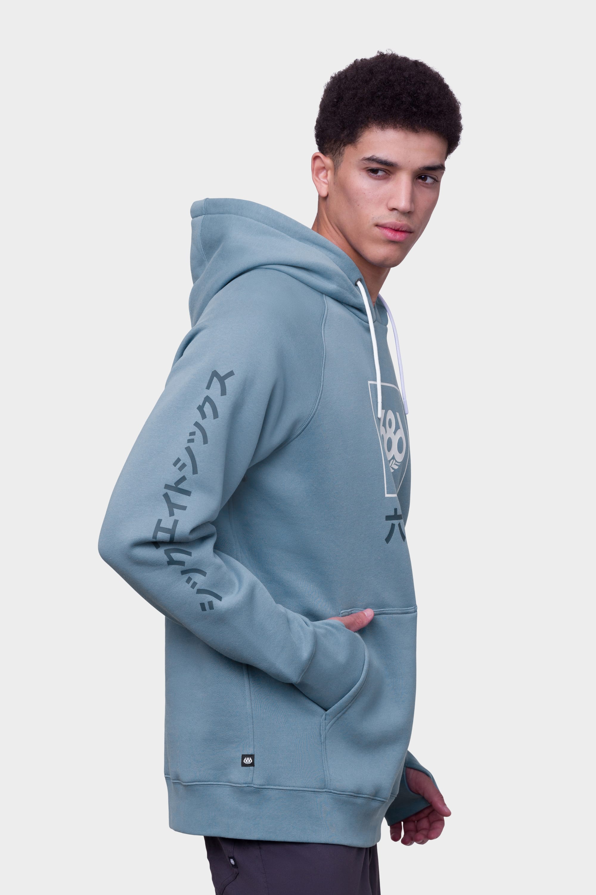 A young man stands sideways against a plain white background, one hand in his pocket, wearing the 686 Mens Script Knockout Hoody—a teal hoodie by 686 featuring white drawstrings, Japanese characters on the sleeve, and a geometric front graphic.