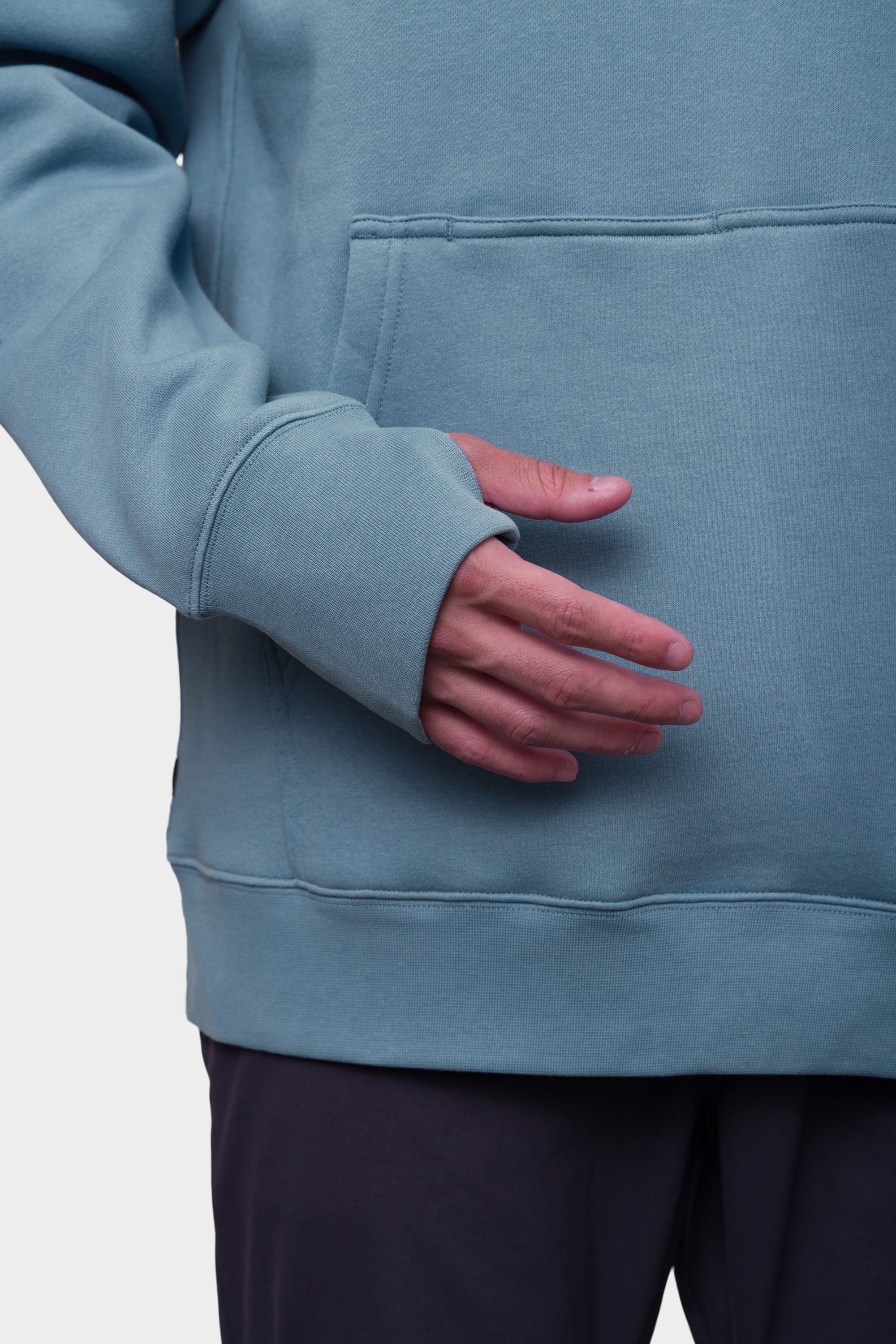 A person wearing the 686 Mens Script Knockout Hoody in light blue, featuring a front pocket and thumb hole sleeves with their hand partially through the opening, paired with dark pants.