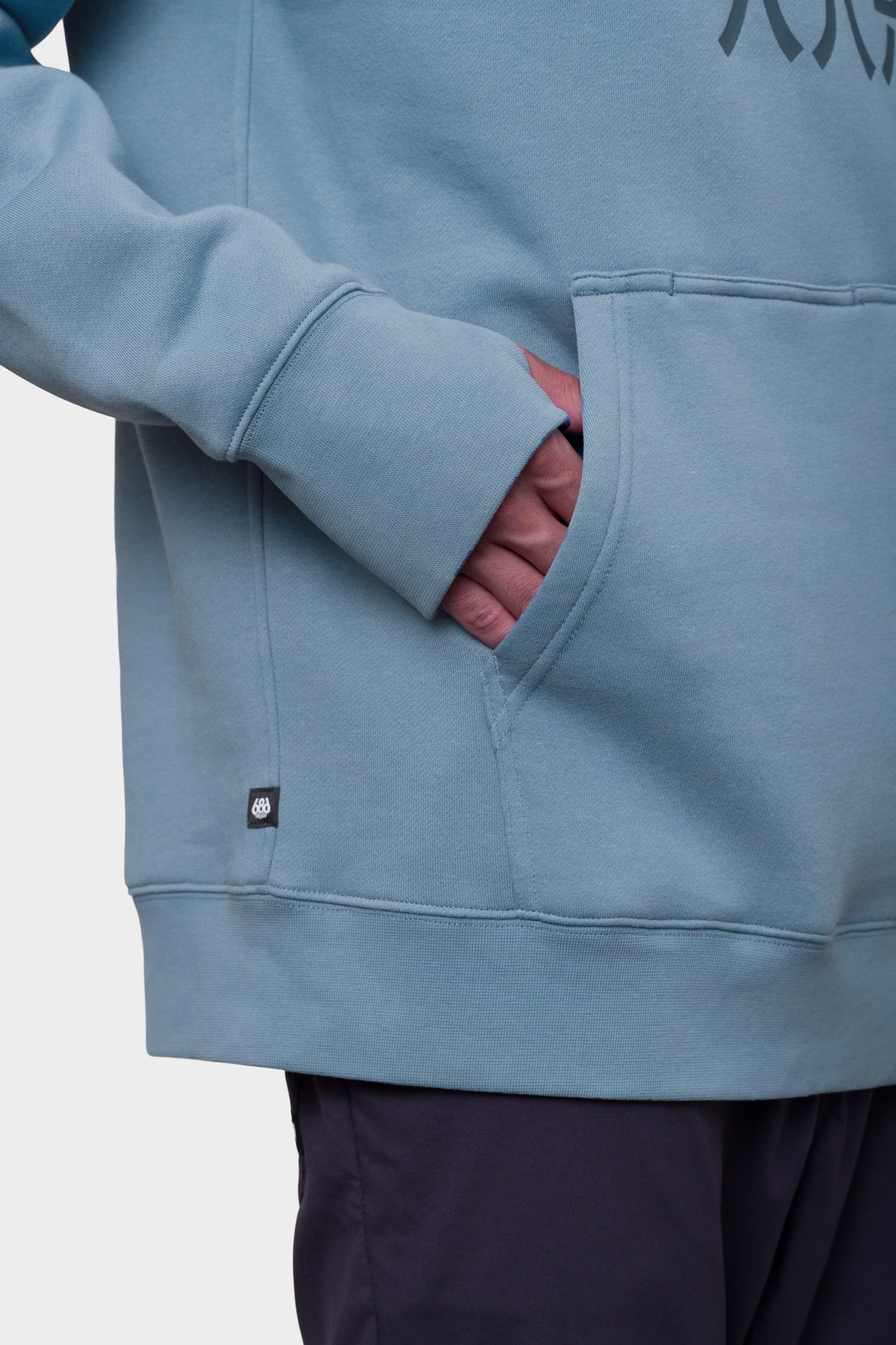 A person is seen from the waist down wearing the 686 Mens Script Knockout Hoody in light blue, with one hand in the front pocket and a plain background behind them. Only the lower torso and part of their arm are visible. Brand: 686.