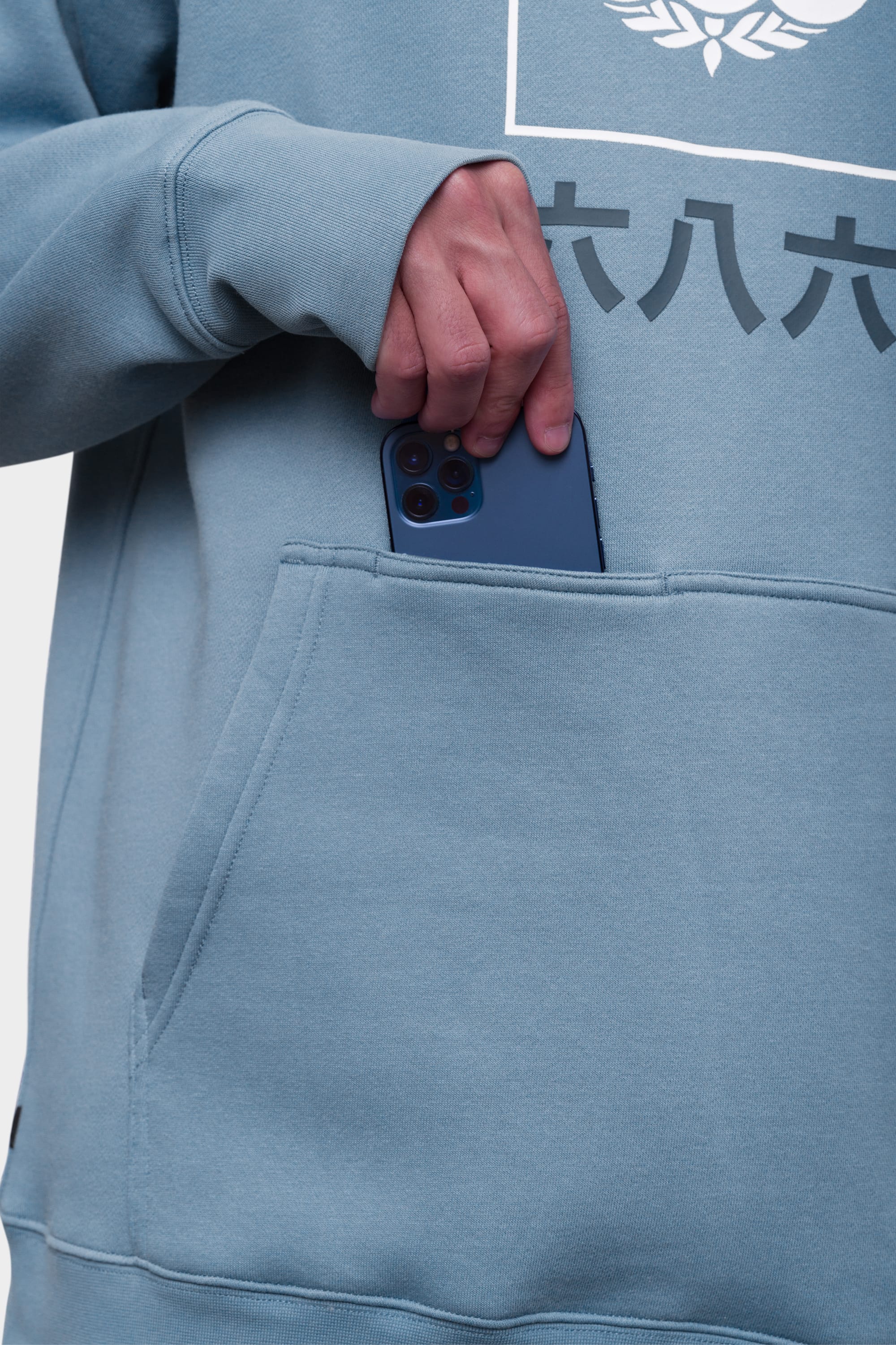 A person wearing the 686 Mens Script Knockout Hoody is placing a blue smartphone into its front pocket. Only their hand and torso are visible. The light blue sweatshirt by 686 features printed text and a design.