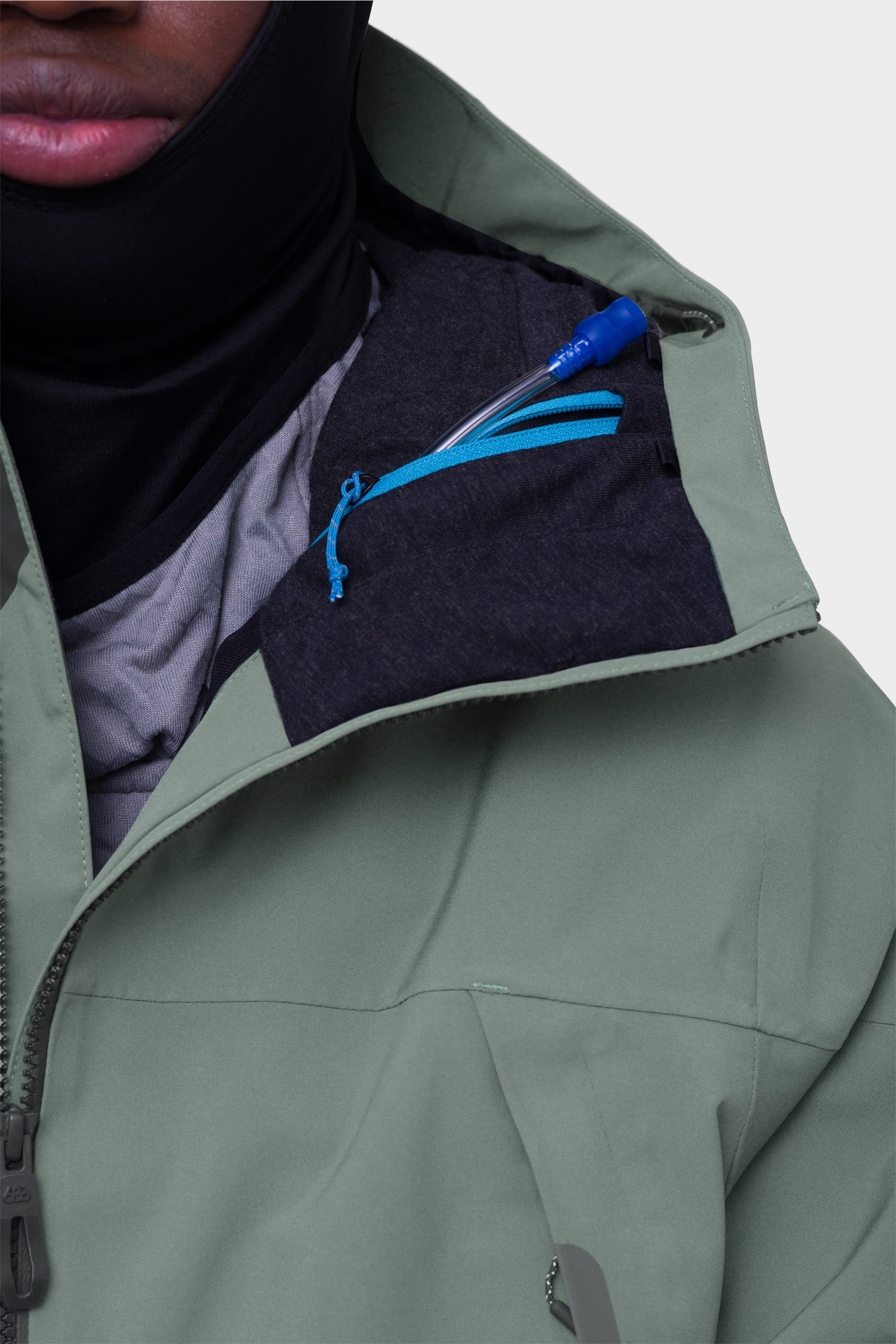 A person wearing the 686 Mens Complete Bladder System by 686, featuring a green jacket with a black balaclava and a blue pen partially visible in the small chest pocket near the collar. Only the lower half of their face is shown.