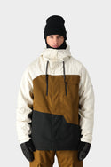 A person in the 686 Mens Geo Insulated Jacket by 686, featuring a geometric pattern in white, brown, and black, pairs it with brown pants, black gloves, and a black beanie while standing against a plain light gray background.