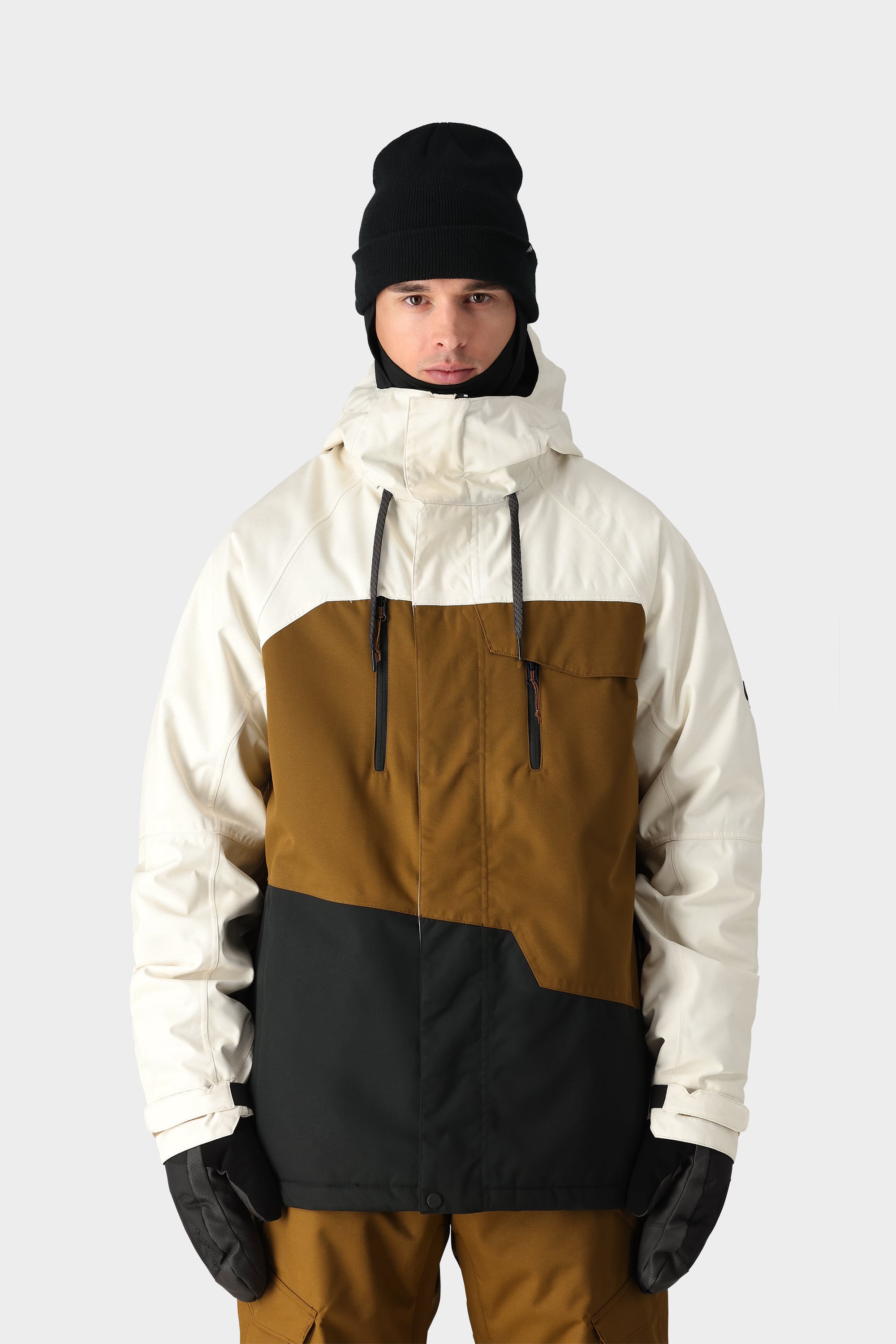 A person in the 686 Mens Geo Insulated Jacket by 686, featuring a geometric pattern in white, brown, and black, pairs it with brown pants, black gloves, and a black beanie while standing against a plain light gray background.