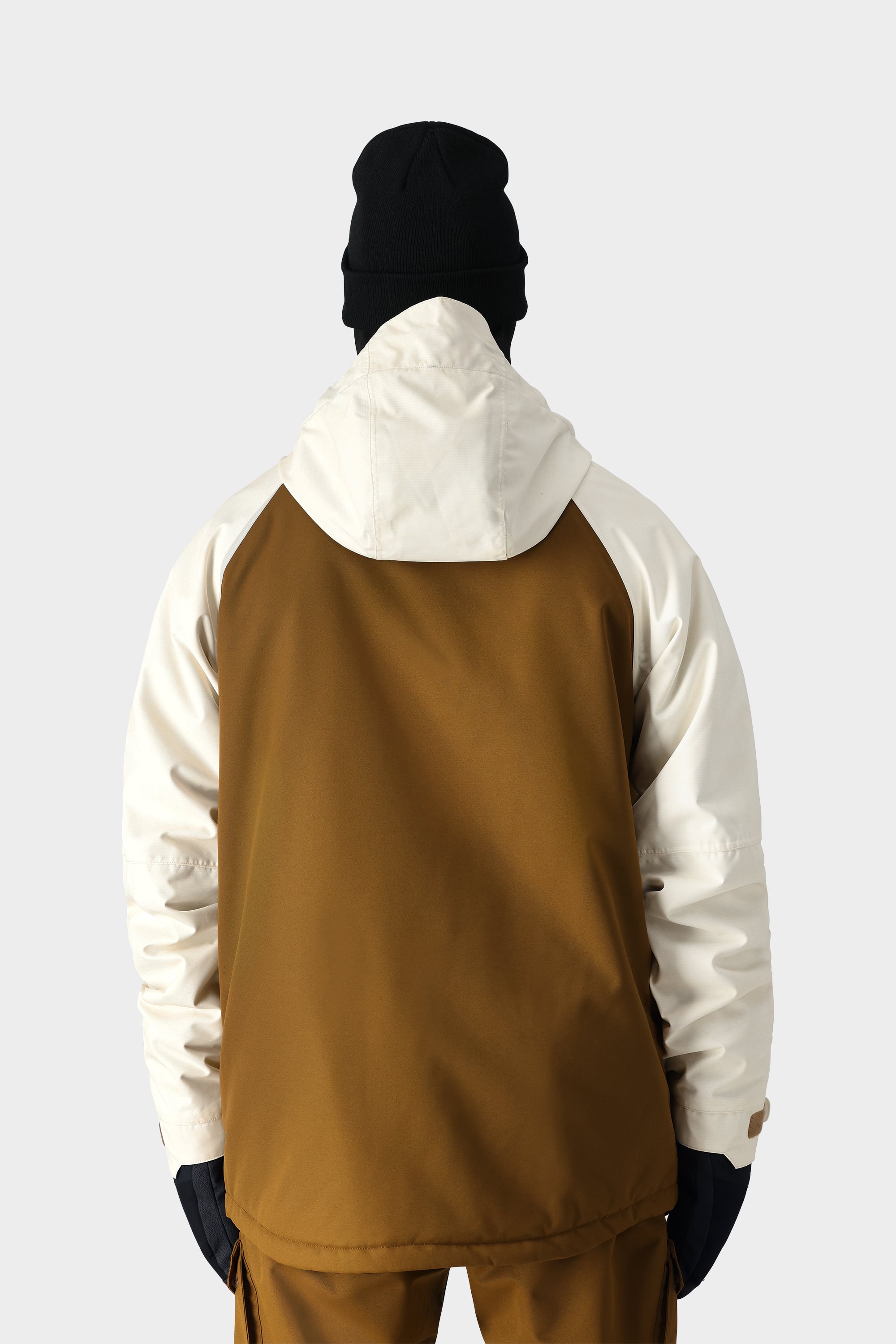 A person wearing the 686 Mens Geo Insulated Jacket in brown and white, along with brown pants, black gloves, and a black beanie, stands with their back to the camera against a plain light gray background.