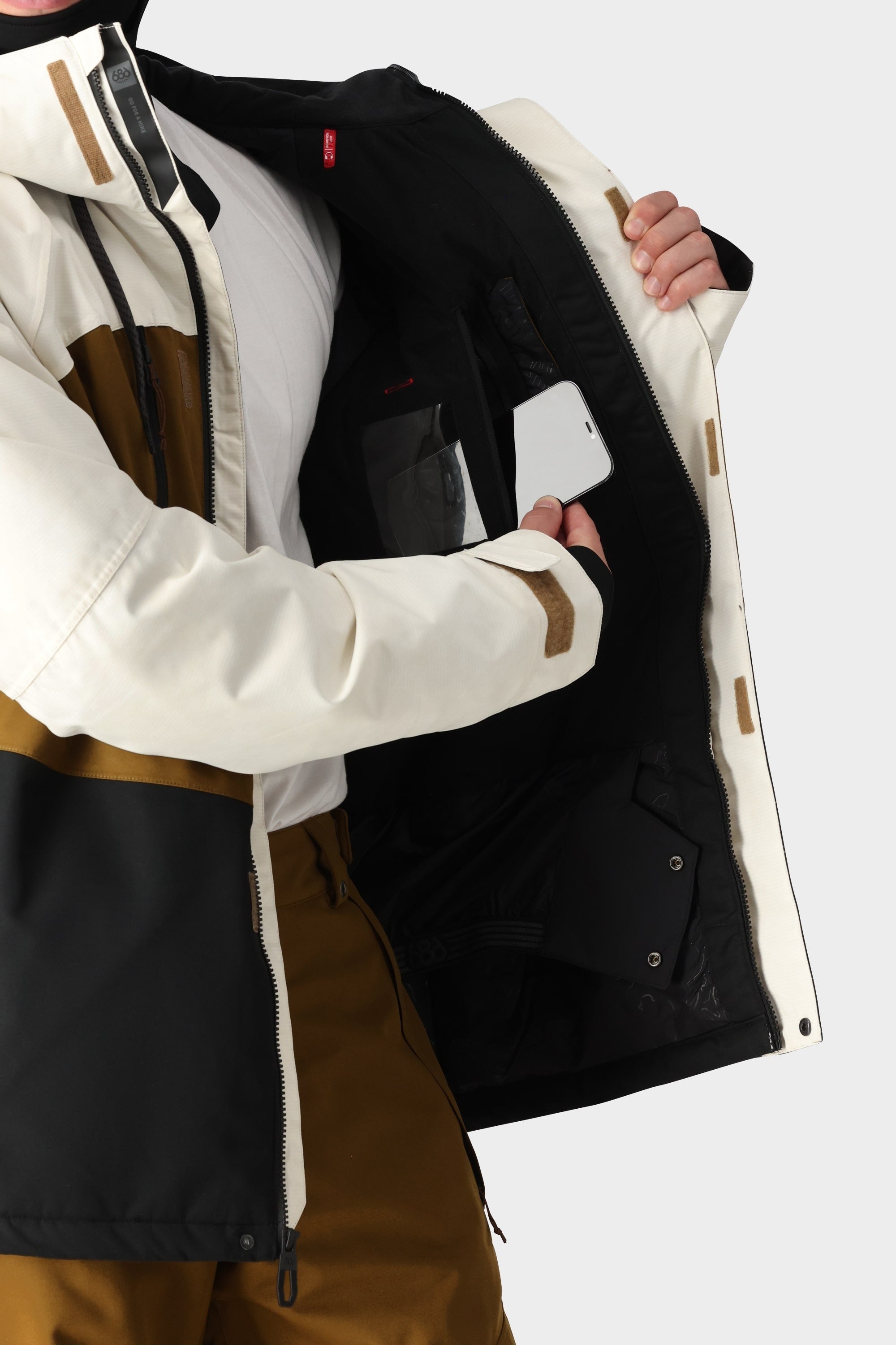 A person wearing the 686 Mens Geo Insulated Jacket in white, brown, and black opens it to show its tech-feature interior pocket, placing a white smartphone inside.