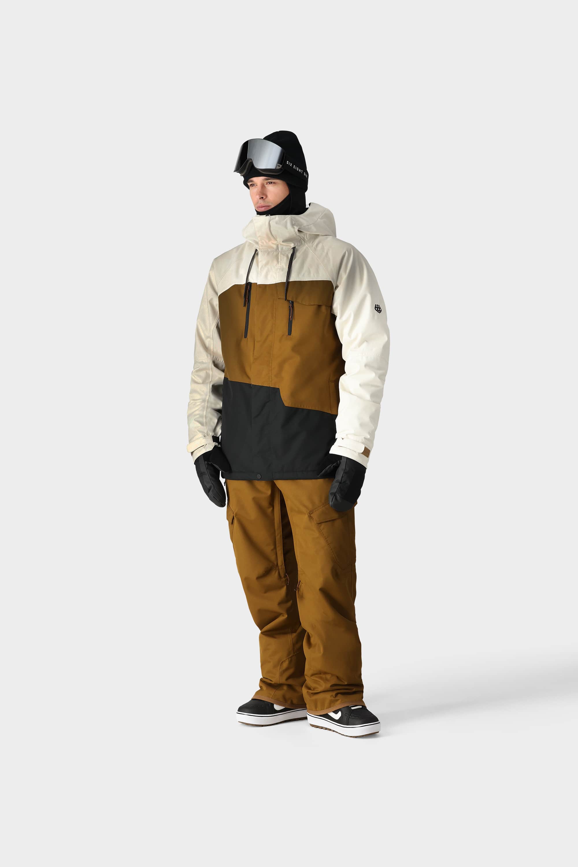 A person wears the 686 Mens Geo Insulated Jacket by 686, featuring a brown and black geometric pattern, paired with black gloves, ski goggles, and black sneakers, standing against a plain light gray background.