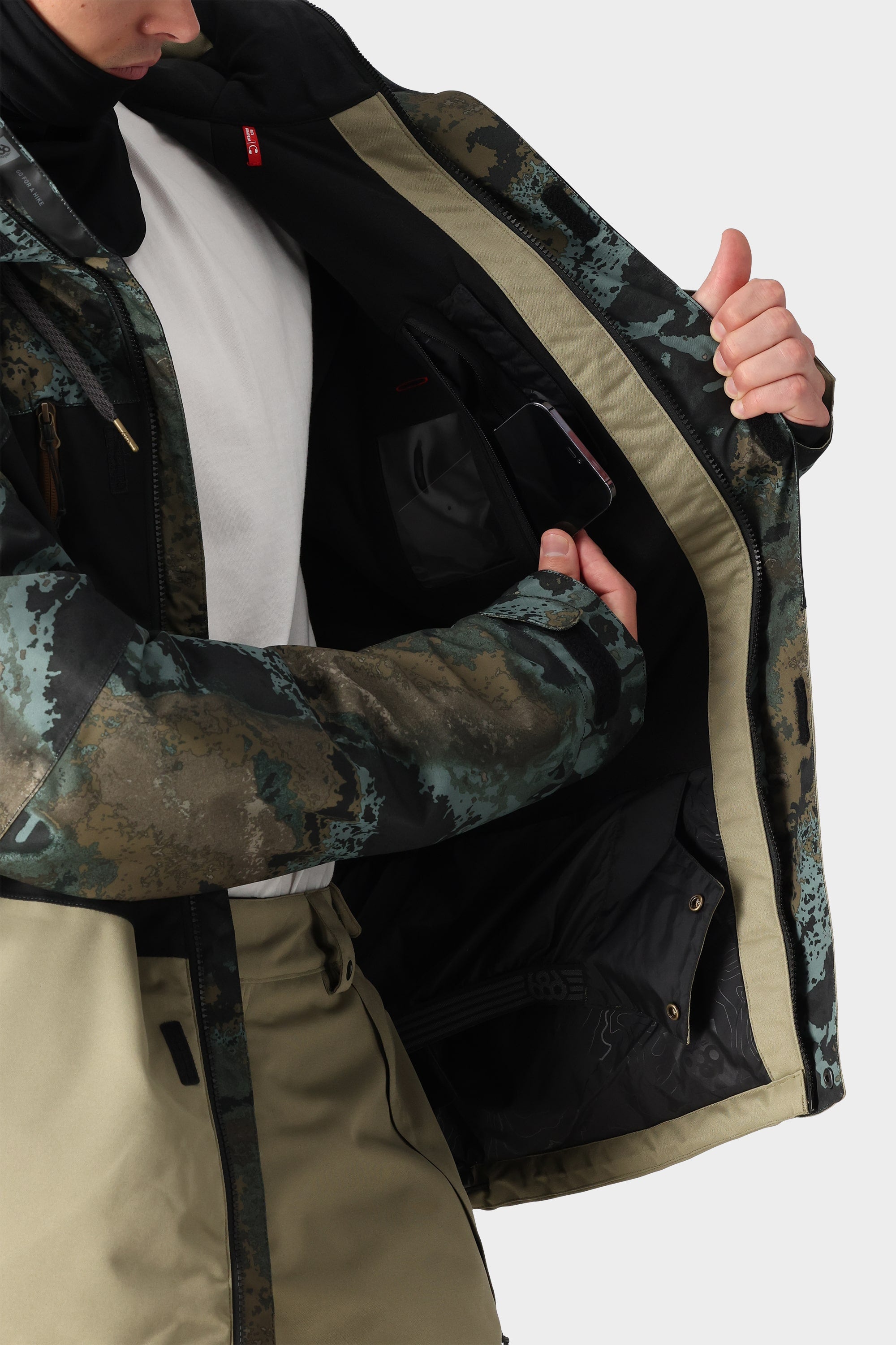 A person in the 686 Mens Geo Insulated Jacket, featuring a patterned exterior and beige lining, opens the front to reveal an inner pocket for their smartphone. Only the torso and hands are visible.