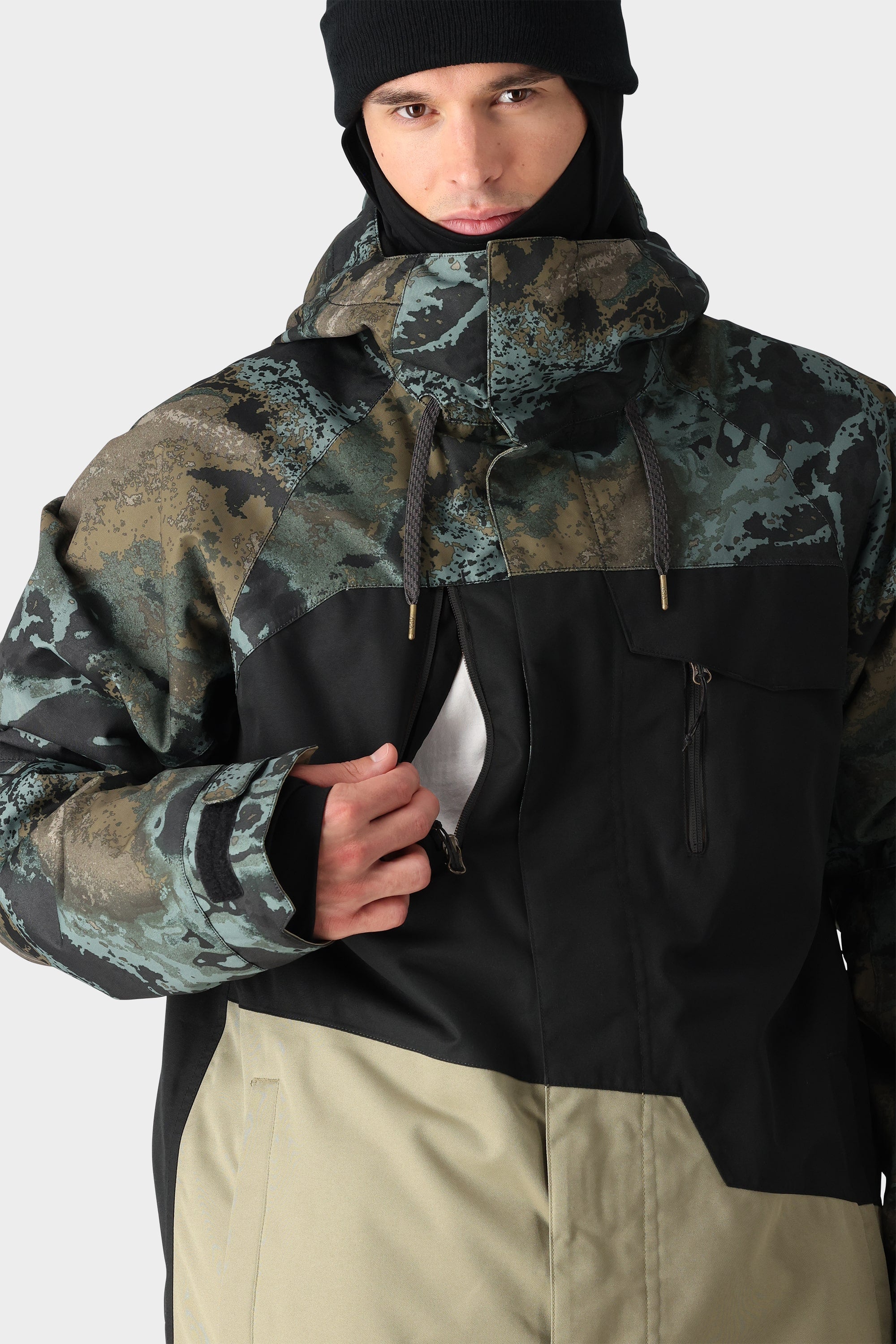 A person in a black beanie is unzipping the chest pocket of the 686 Mens Geo Insulated Jacket, featuring a geometric camouflage pattern with dark green, black, and beige color blocks and a high collar. Brand: 686.