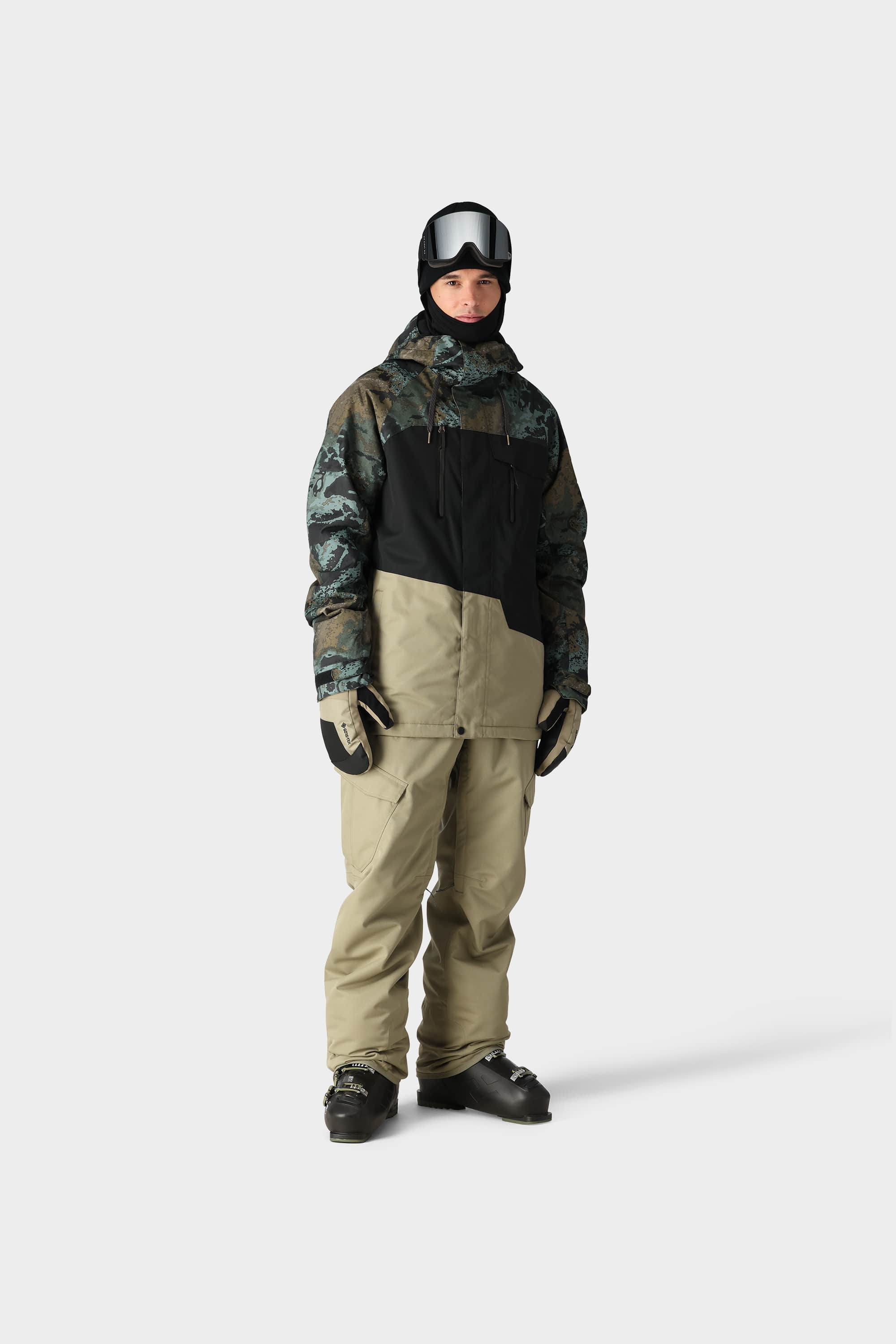 A person stands against a plain light background in full snowboarding gear, wearing the 686 Mens Geo Insulated Jacket by 686 with black gloves, beige pants, ski goggles, and a helmet.