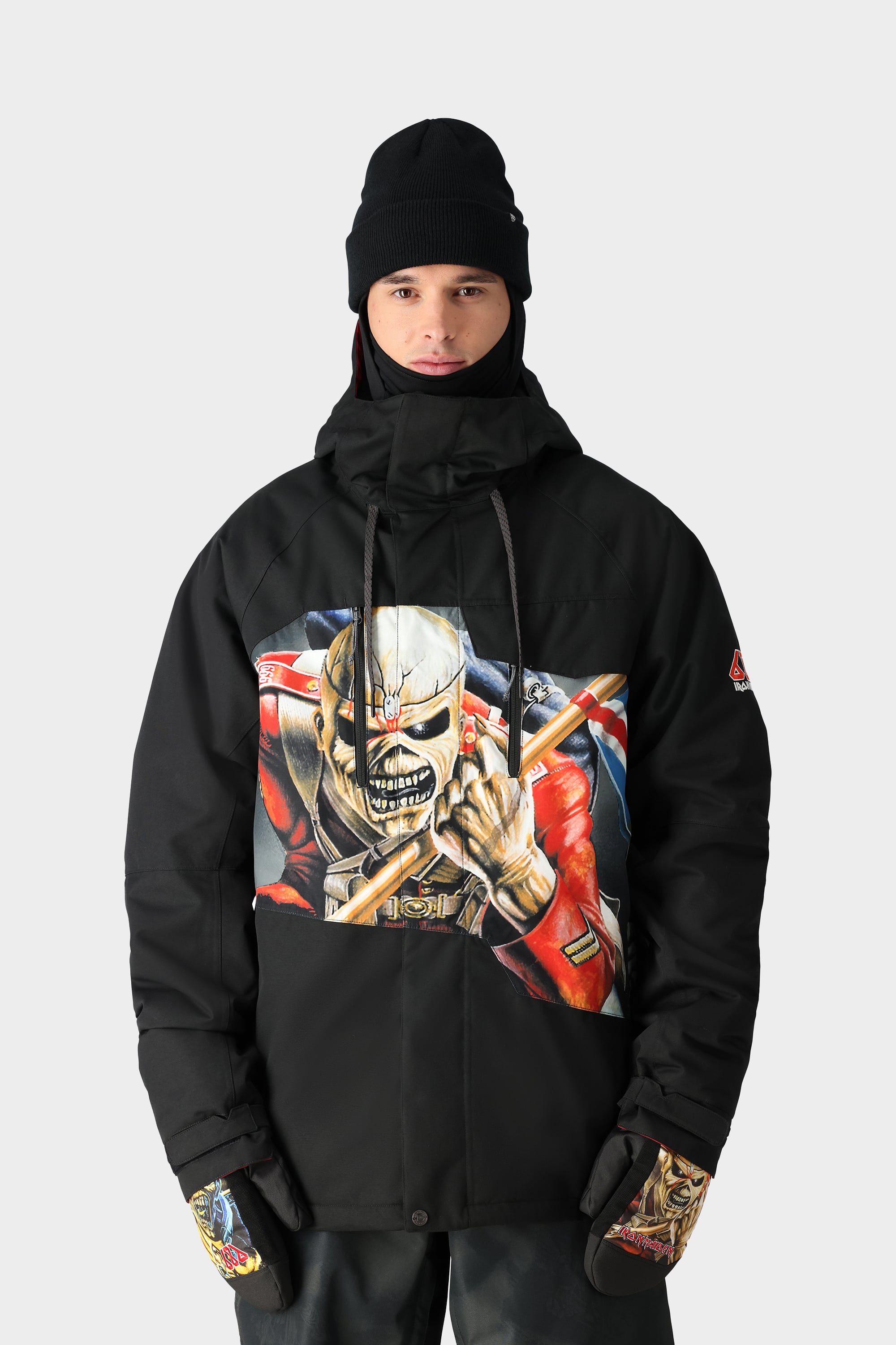 A person in the 686 Mens Geo Insulated Jacket, which features a bold skeleton graphic with a red jacket and axes, plus a black beanie, gloves, and dark pants from 686, stands against a plain light background.