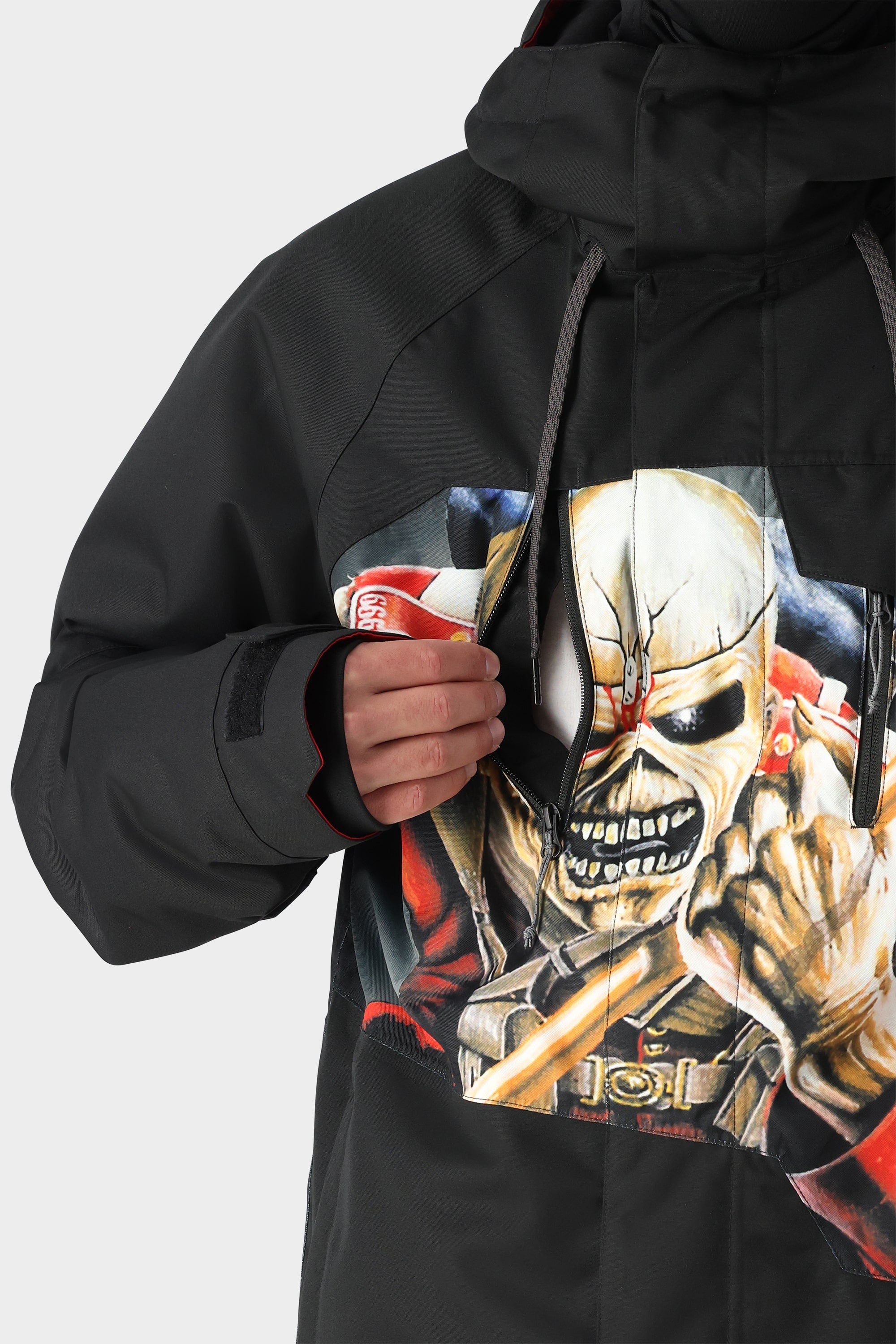 A person wearing the 686 Mens Geo Insulated Jacket in black, featuring a bold graphic of a menacing skeletal figure, partially unzips a front pocket on this affordable 686 brand jacket.