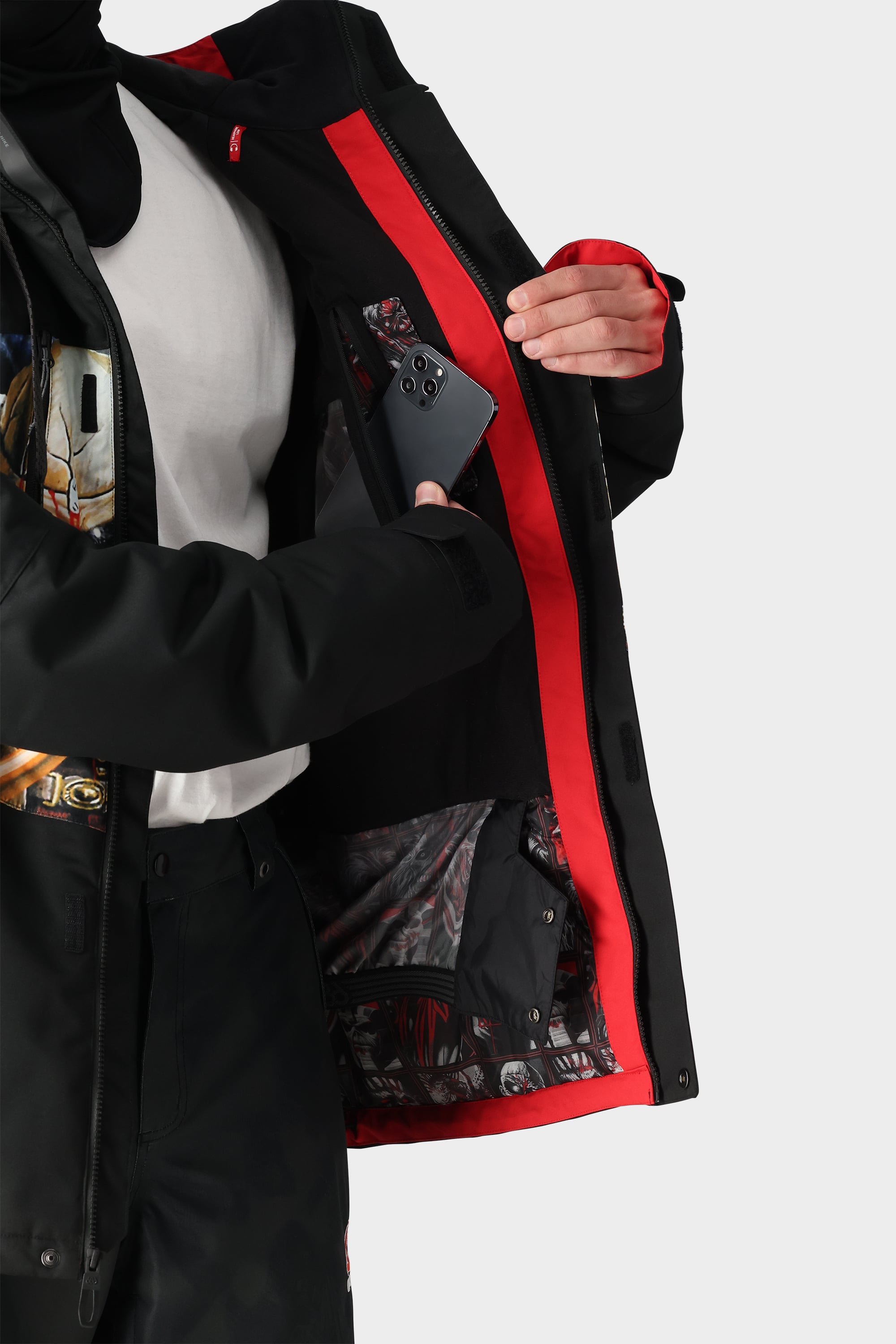 Wearing the 686 Mens Geo Insulated Jacket with tech features, a person puts a smartphone into an inner pocket, showcasing the bold patterned lining. A white shirt with a visible colorful graphic is worn underneath.