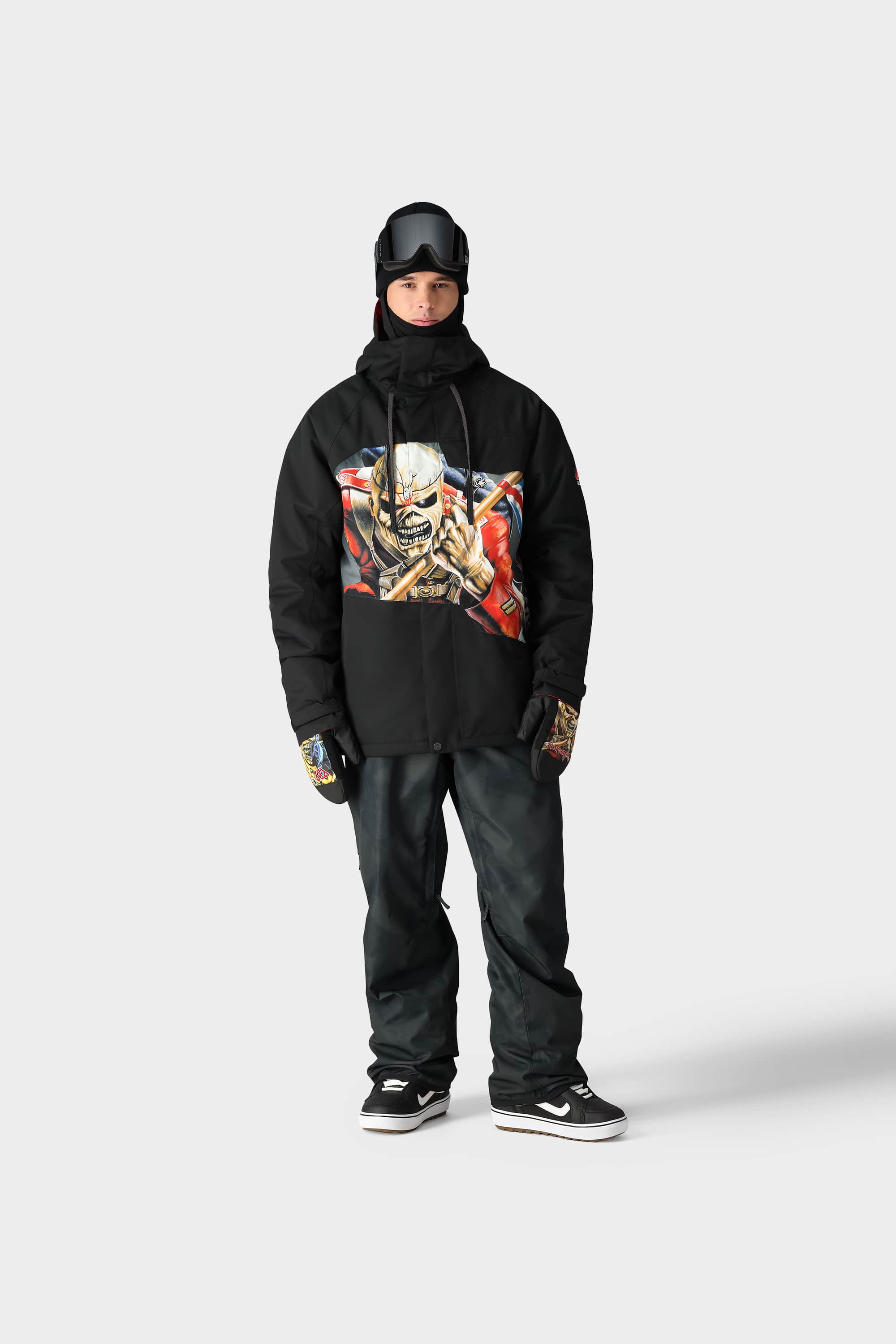 A person in black snowboarding gear, featuring the 686 Mens Geo Insulated Jacket by 686 with a graphic screaming face, black pants, gloves, goggles, and sneakers, stands against a plain white background.