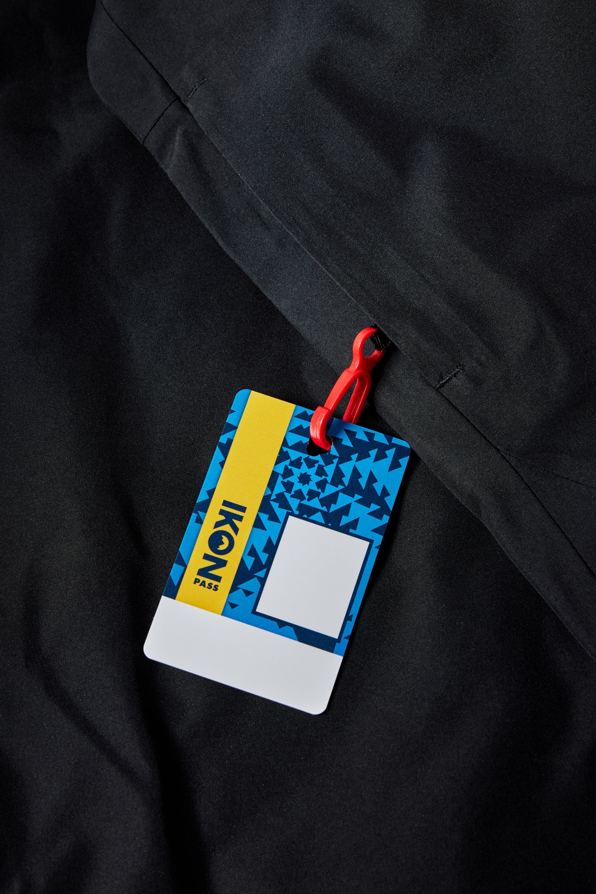 A colorful Ikon Pass ski lift card with blue, yellow, and white geometric patterns is attached to a red clip, resting on black fabric—perfect to pair with your 686 Mens Geo Insulated Jacket from 686.