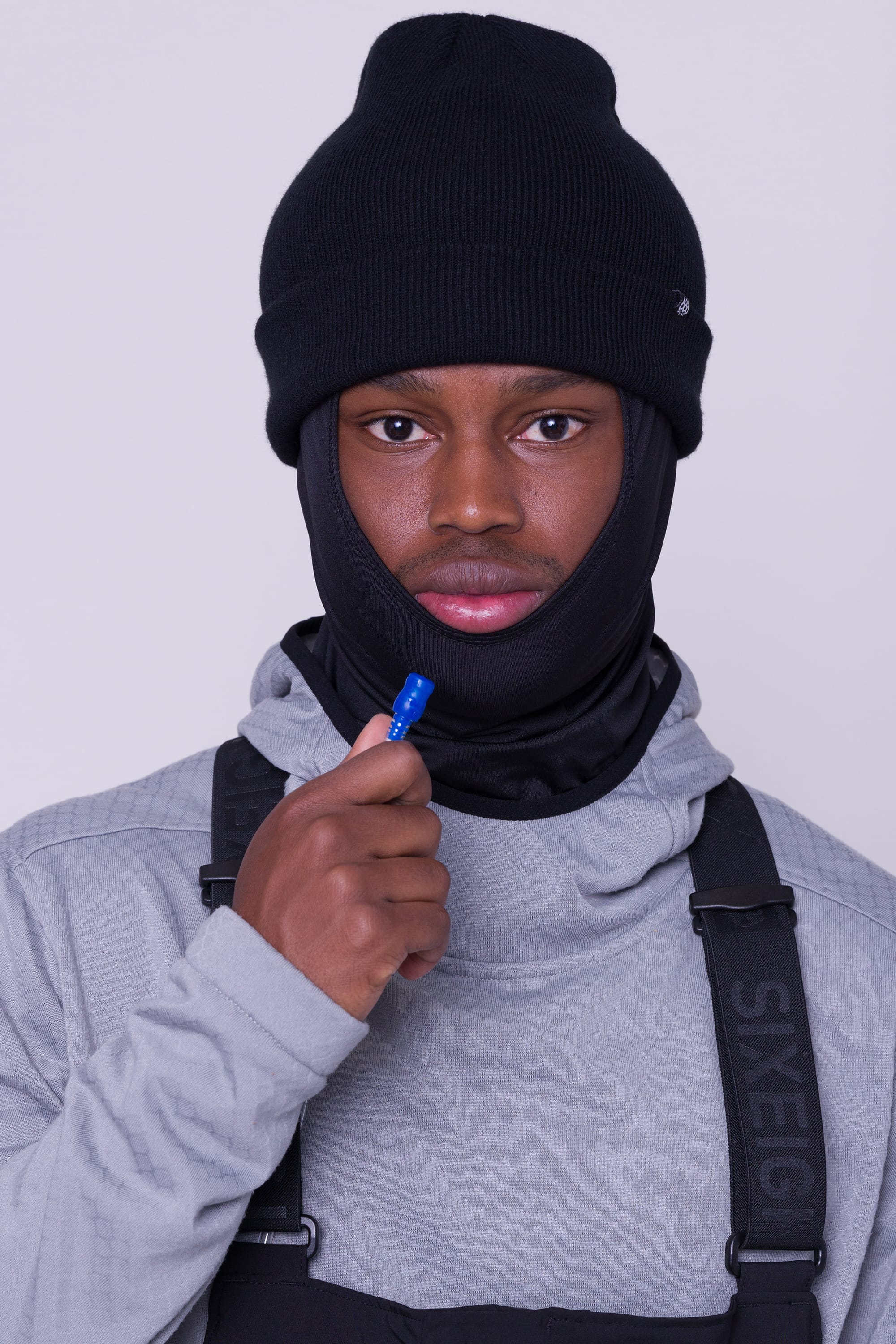 A person in a gray hoodie, black beanie, and balaclava wears the 686 Mens Complete Bladder System by 686, holding its blue hydration tube near their mouth and looking directly at the camera.